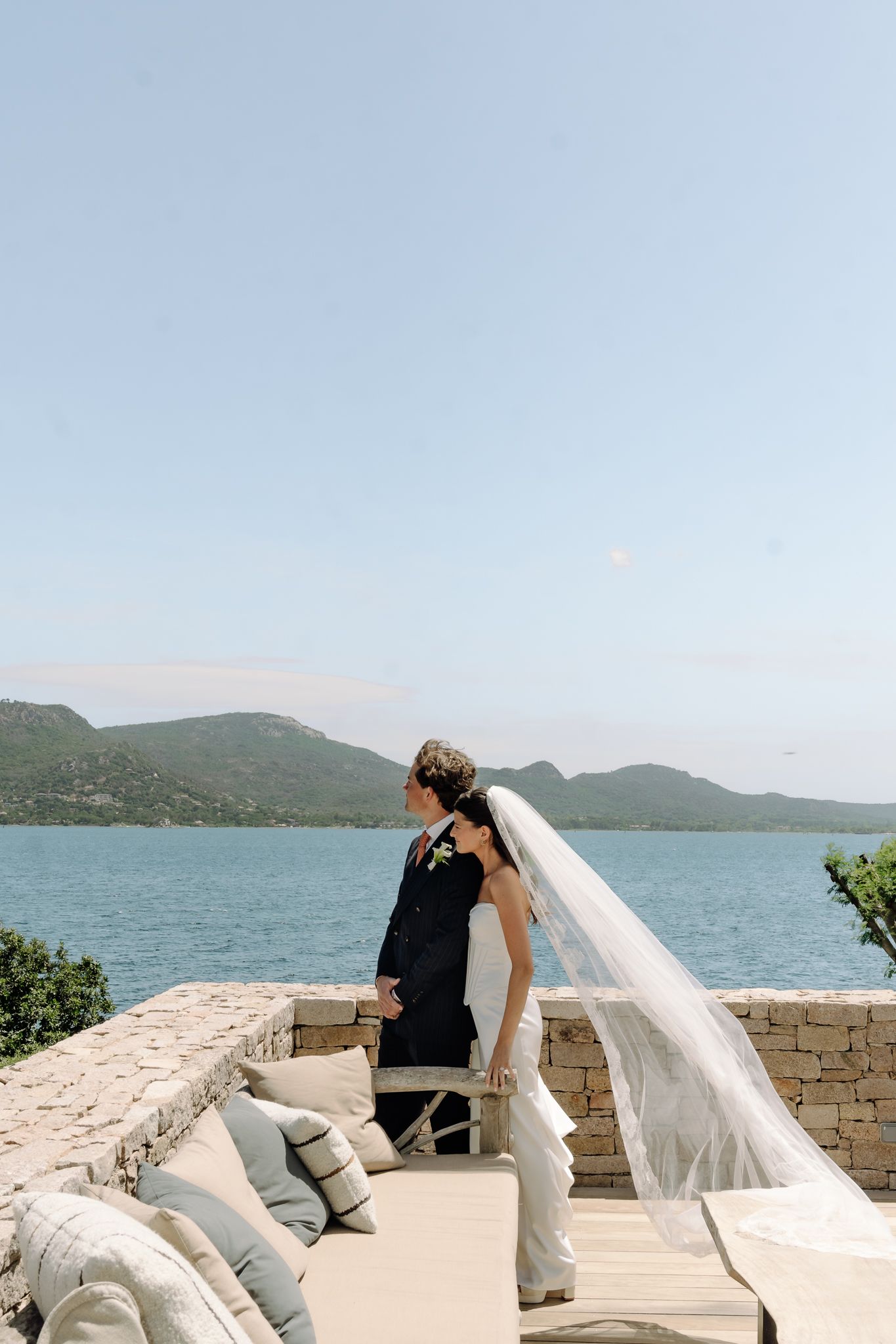 Newlyweds stand together on a coastal terrace overlooking a blue bay and green mountains on a sunny wedding day.