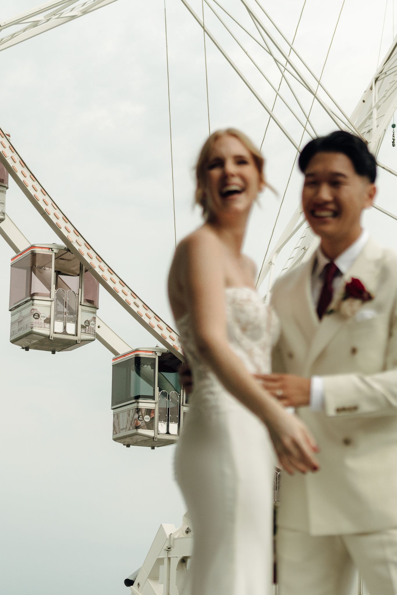 Laughing bride and groom in wedding attire pose in front of a large Ferris wheel on their wedding day.
