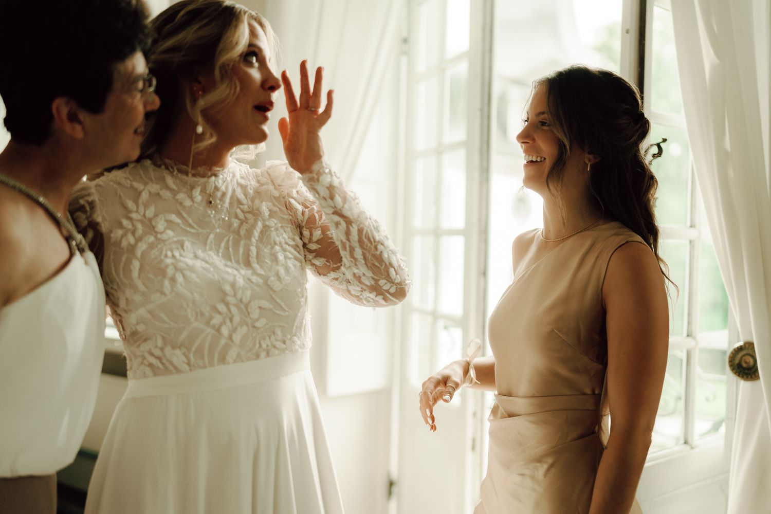 Bride in lace wedding dress laughing and gesturing with bridesmaids near bright windows.