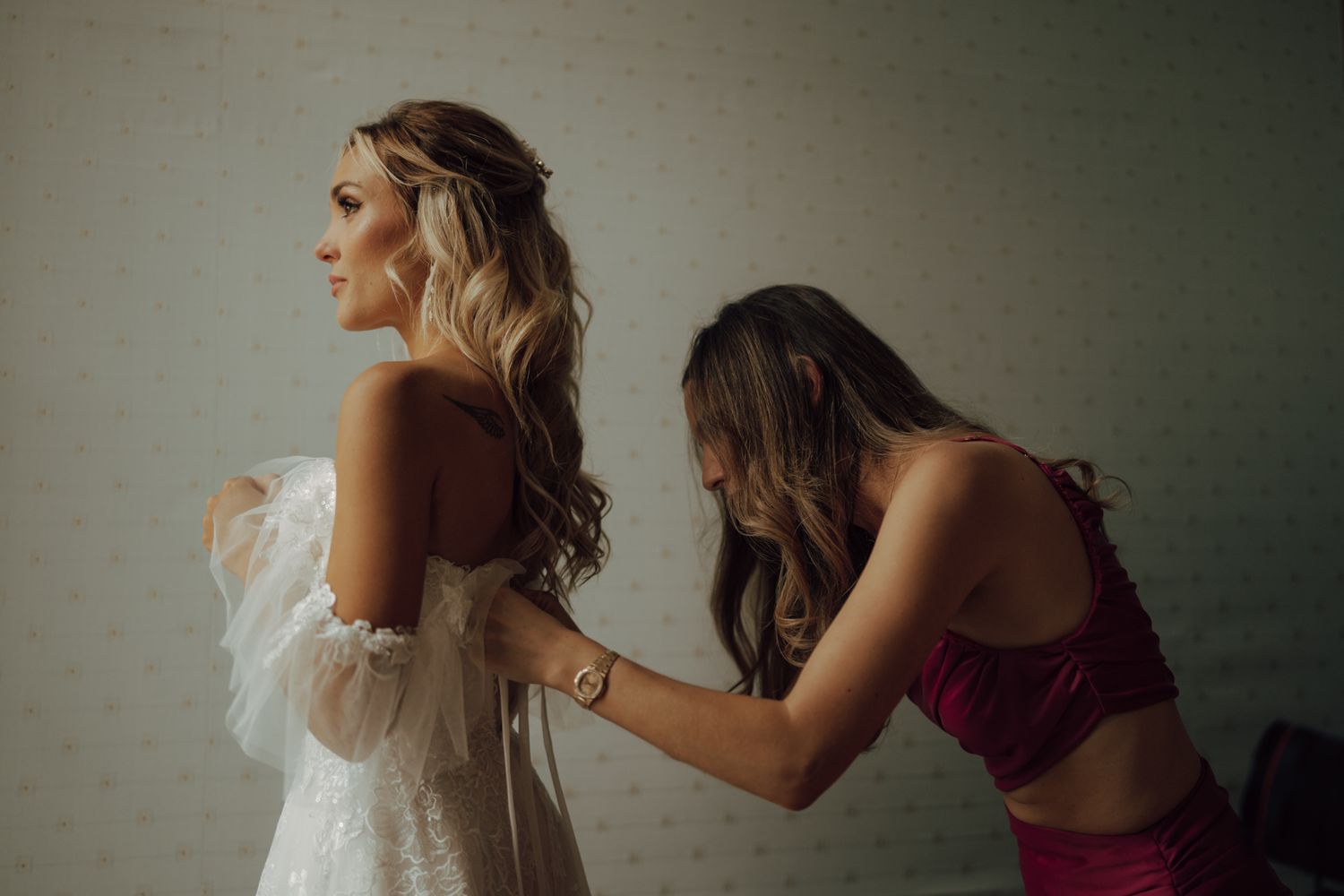 Bridesmaid in red helps bride into strapless embellished wedding gown from behind.