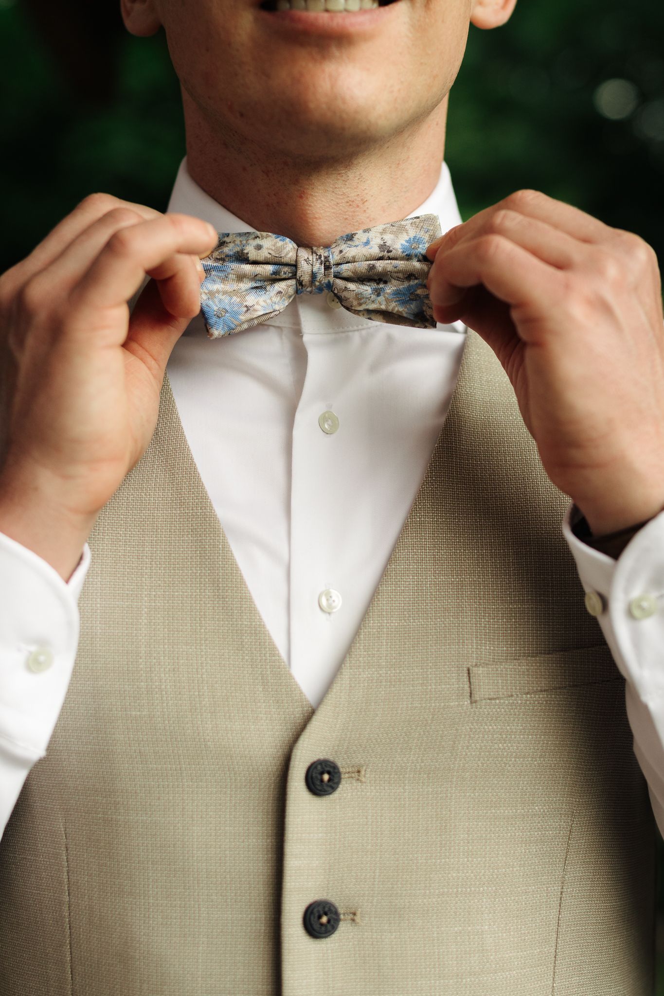 Smiling groom adjusting floral bow tie on beige wedding vest.