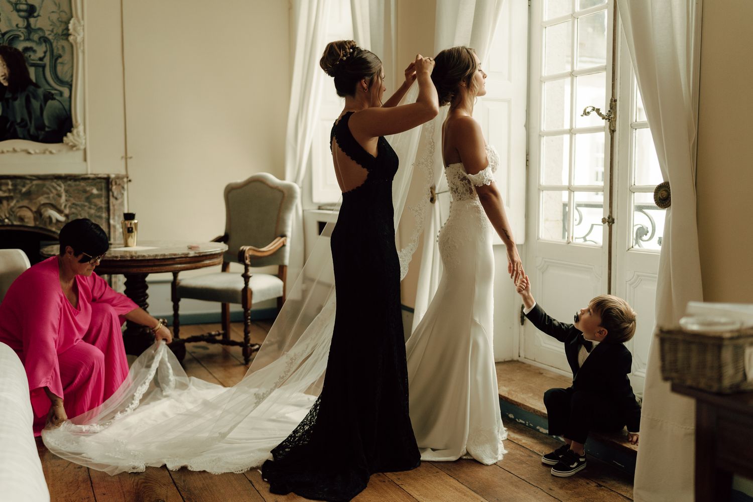Bridesmaids helping bride into white wedding dress in a bright French chateau room.