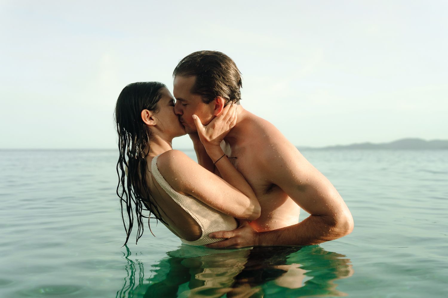 Romantic couple kissing in calm ocean waters, man with tattoos embracing woman with wet hair.