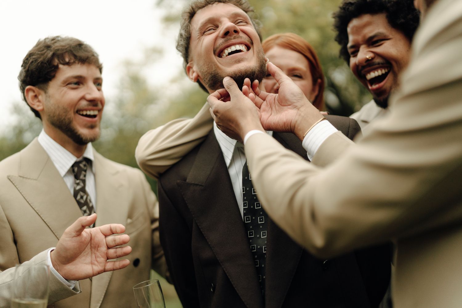 Group of smiling businessmen in suits laughing and adjusting colleague's tie outdoors at a corporate event.