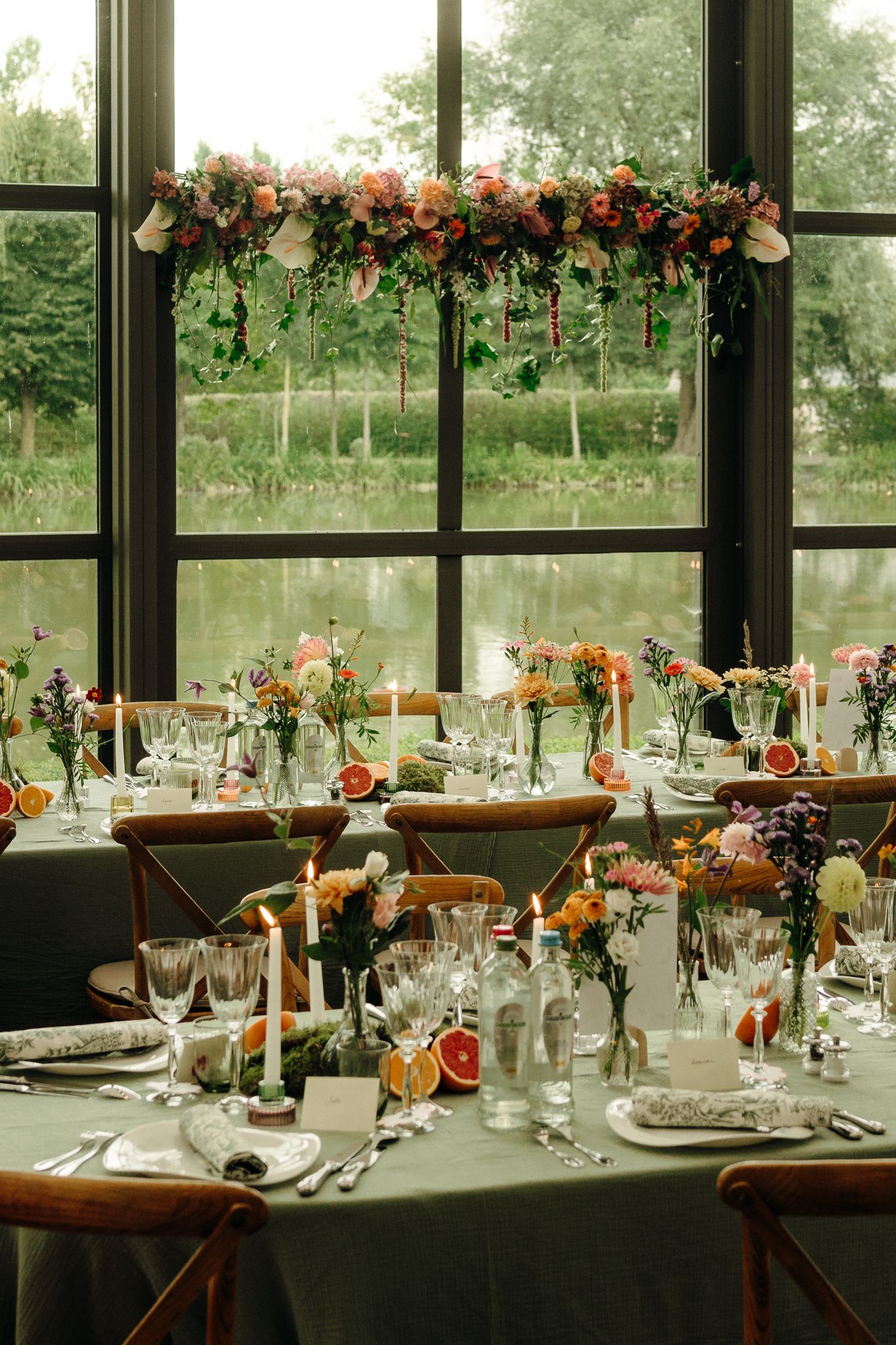 Elegant wedding reception table with green linen, candles, colorful florals, and a floral chandelier by a large window.