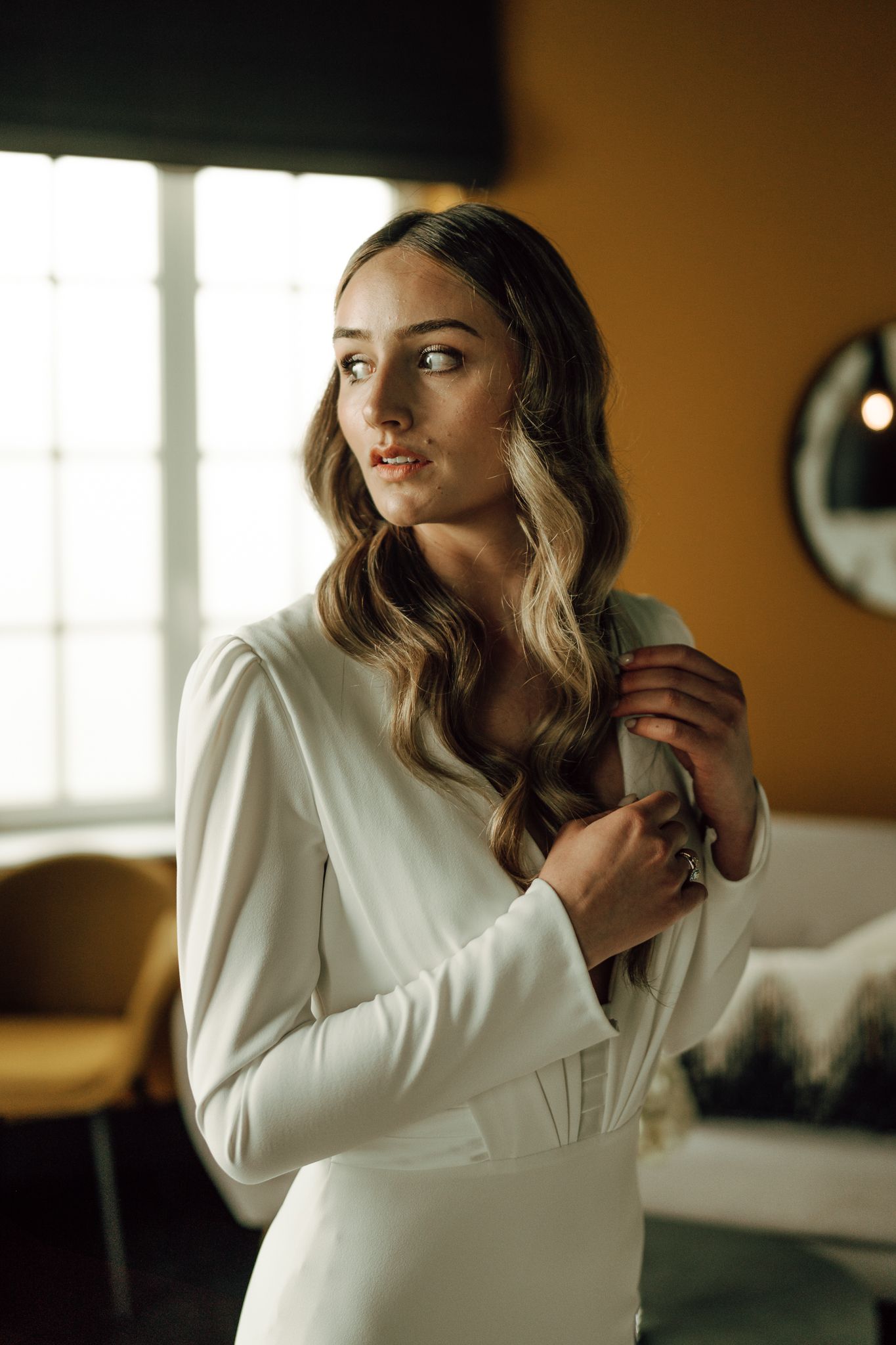 Elegant bride in a long-sleeve white wedding dress adjusting her collar indoors in warm light.