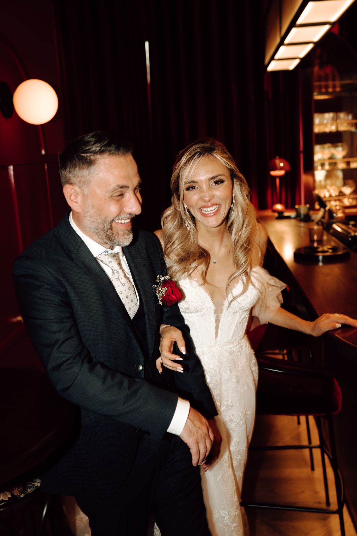 Bride and groom laughing together in warmly lit bar setting during wedding reception.