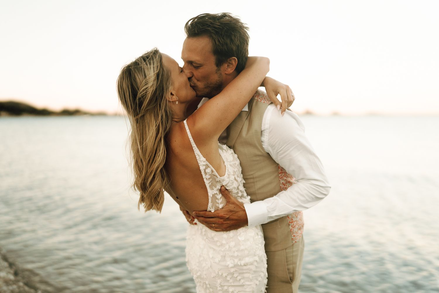Bride and groom embrace and kiss near shimmering water at golden hour.