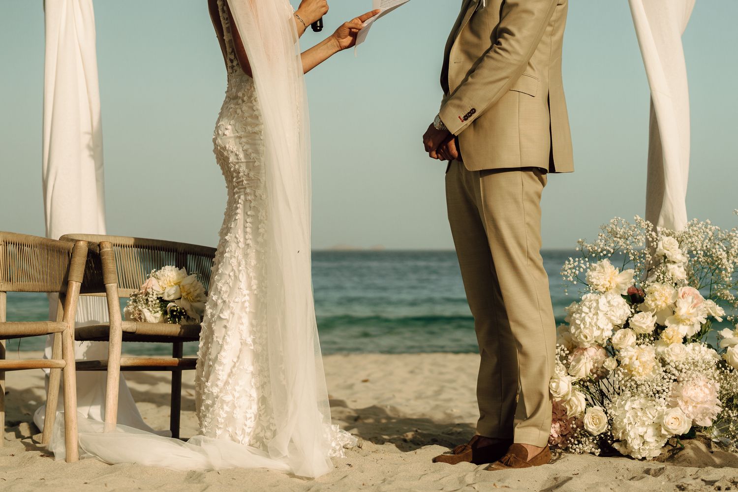 Bride and groom stand barefoot on beach during wedding ceremony beside white floral arrangements.