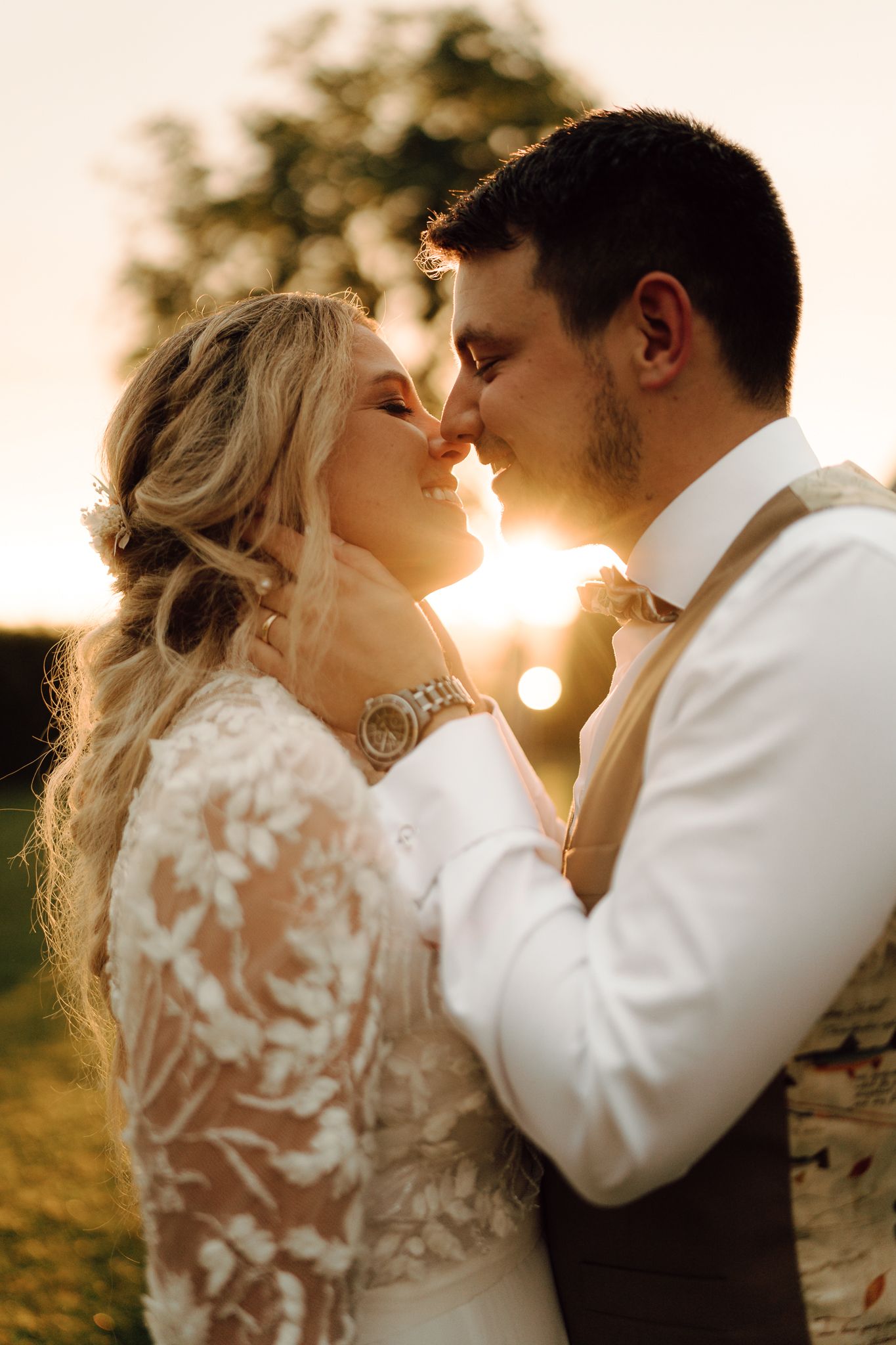 Bride and groom sharing romantic almost-kiss moment bathed in golden sunset backlight outdoors.
