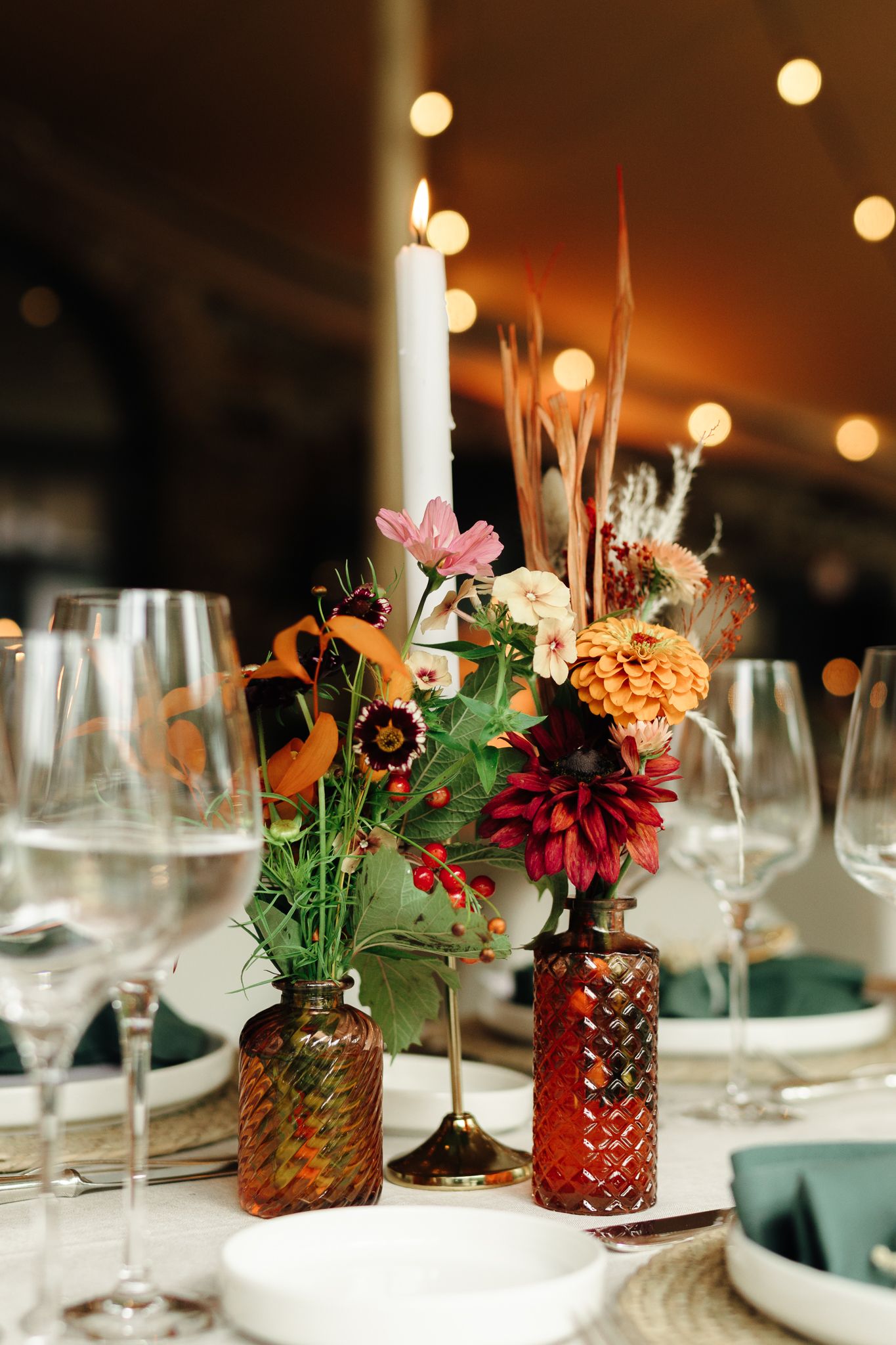 Colorful floral centerpiece with dahlias and wildflowers beside a white candle at wedding table.