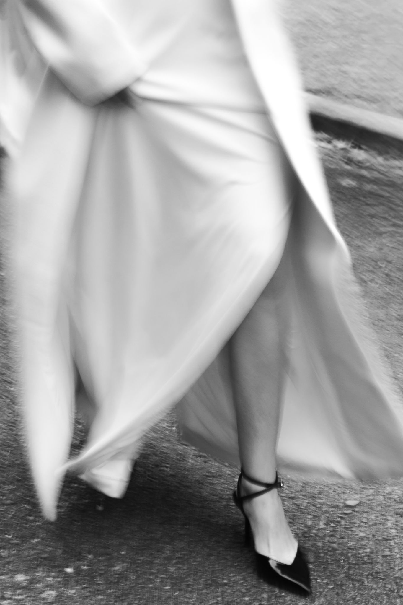 Black and white photo of a woman in a flowing white gown and black heels walking gracefully on a street.