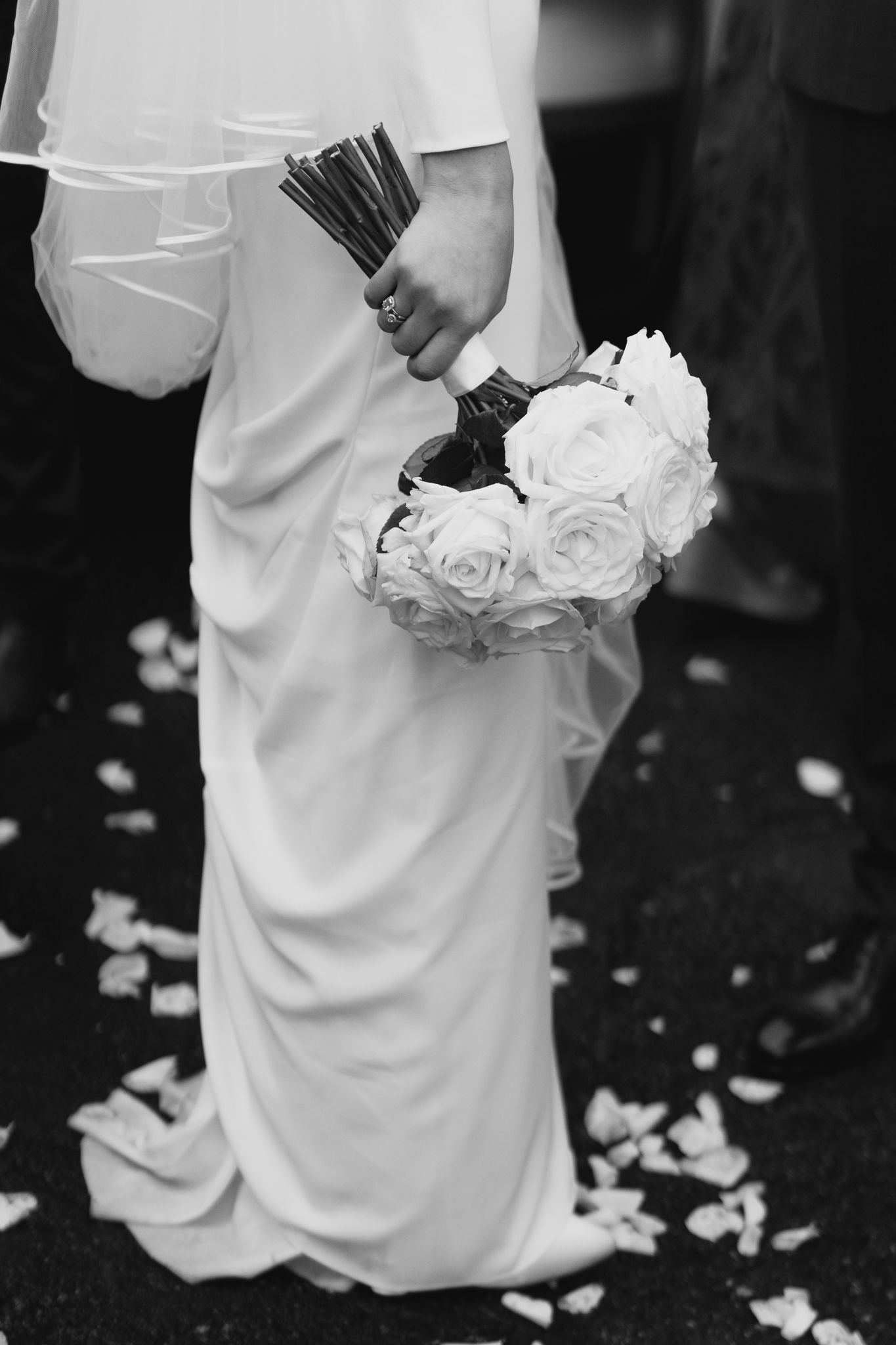 Black and white photo of bride holding a bouquet of white roses with petals scattered on ground.