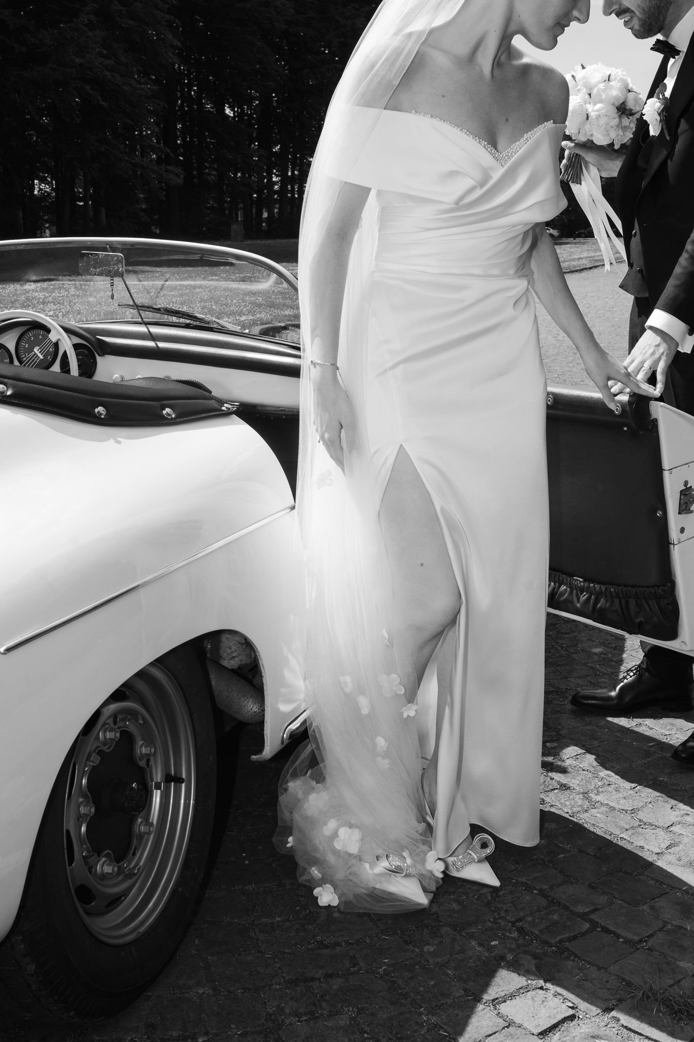 Black and white photo of bride in slit satin gown stepping out of a vintage white convertible.