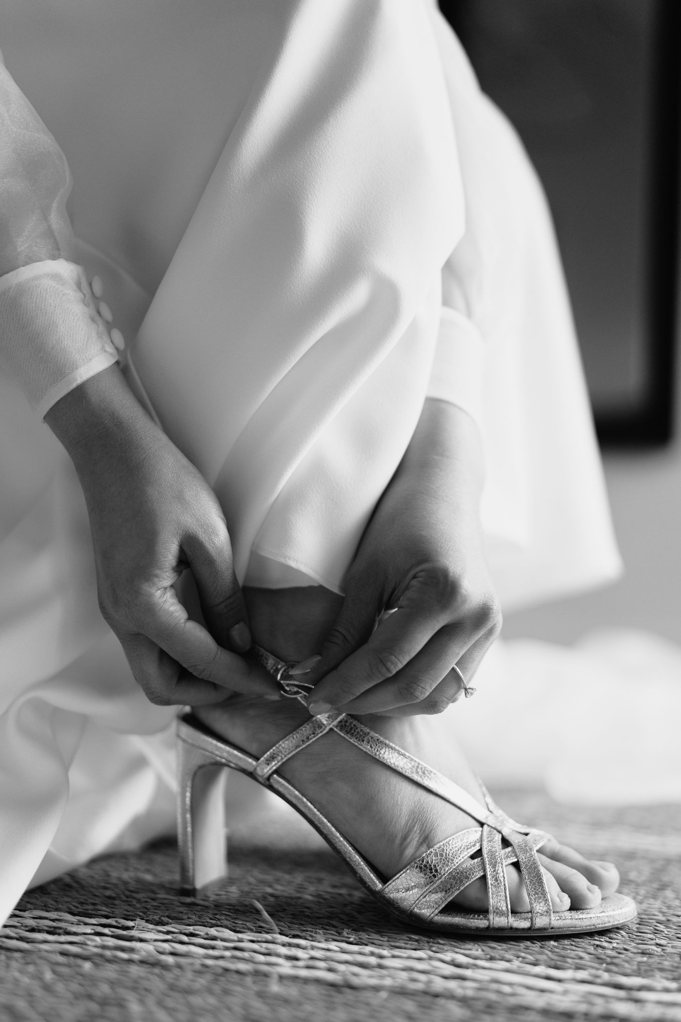 Black and white close-up of bride fastening strappy silver heeled sandal beneath white wedding dress.