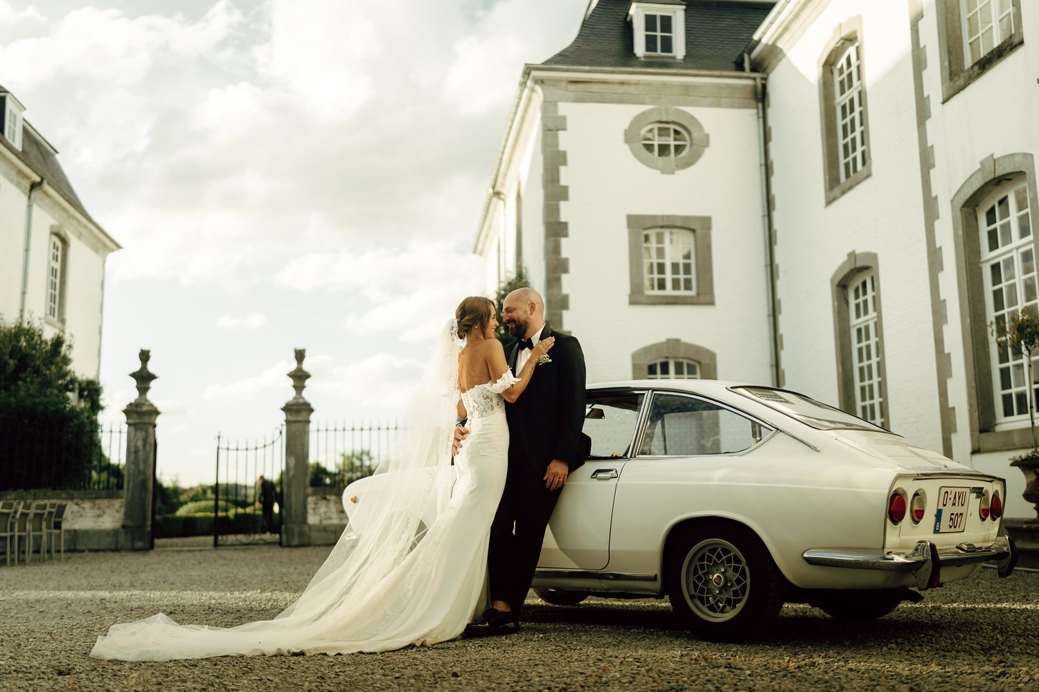 Bride and groom embrace beside a vintage white classic car outside a stunning European chateau wedding venue.