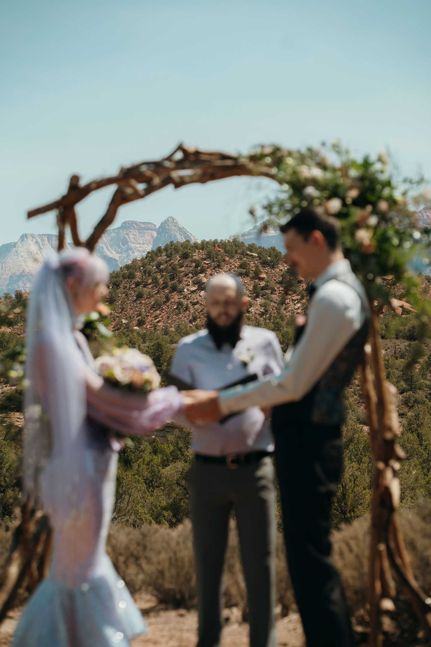 Outdoor wedding ceremony with couple exchanging vows under rustic wooden arch, mountains and desert landscape in background.
