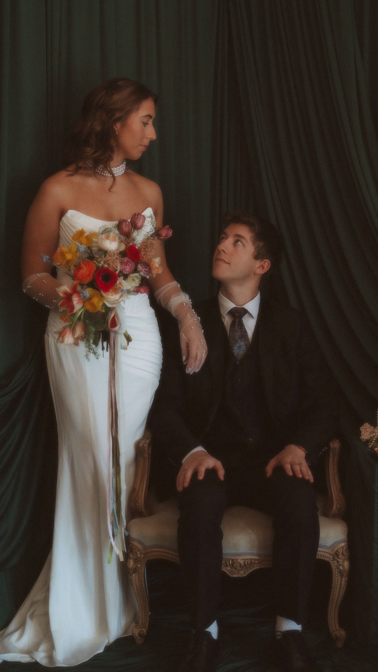 Bride in white mermaid gown with colorful bouquet stands beside seated groom before green drapes.