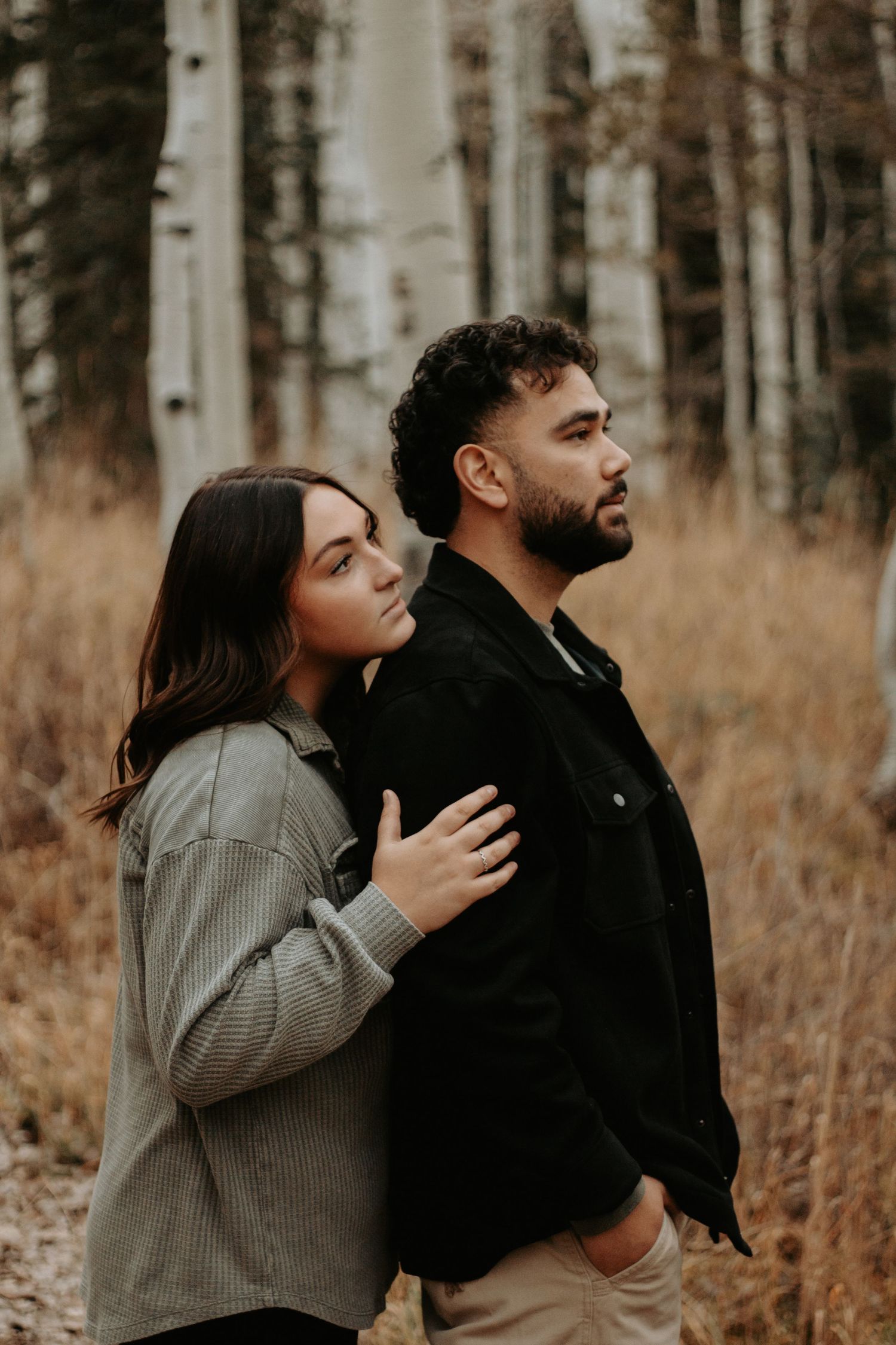 Couple embracing in autumn forest with tall aspen trees, woman in olive jacket resting hands on mans black shirt.