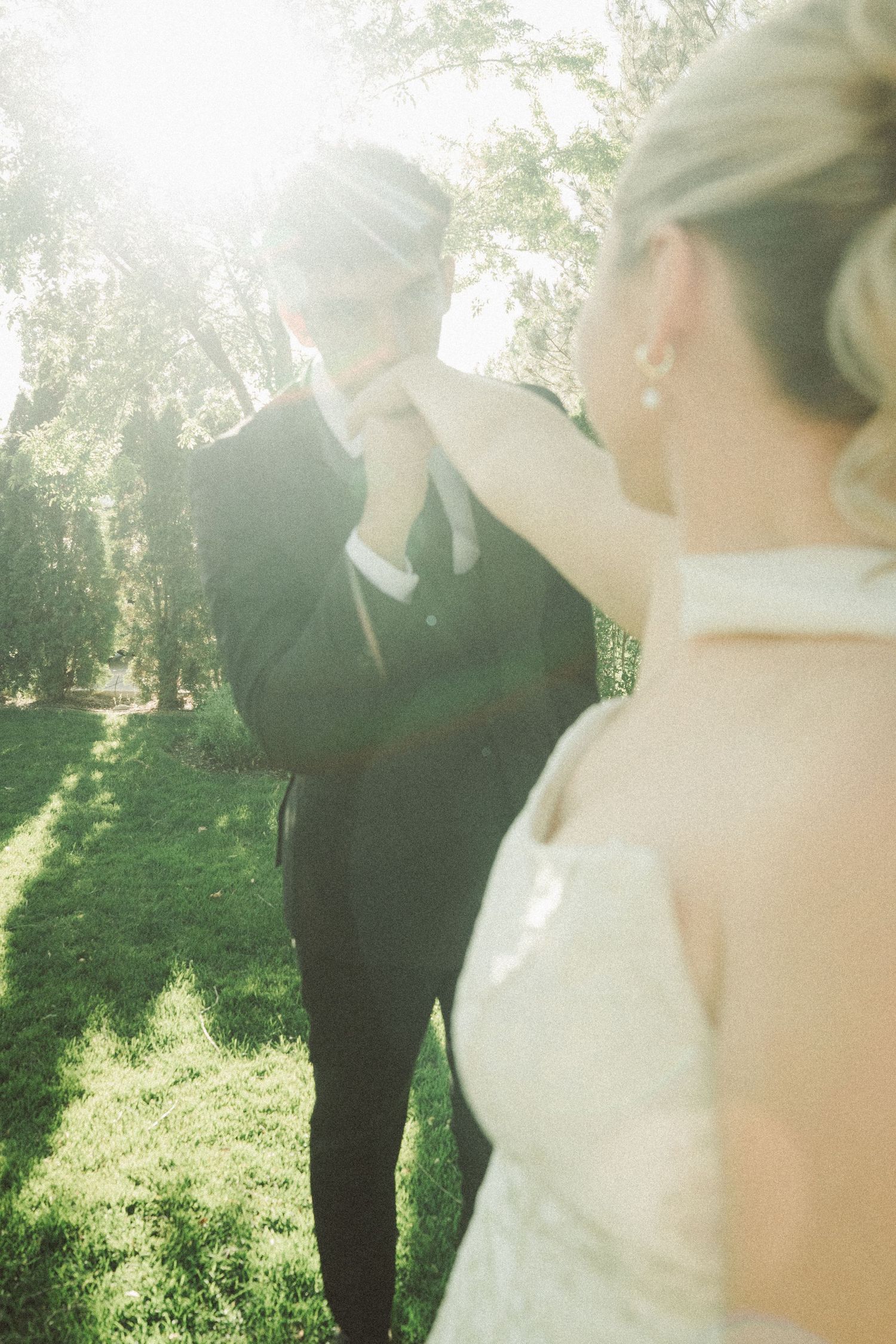 Emotional groom covering his face during outdoor wedding first look with bride in white dress surrounded by lush green trees.