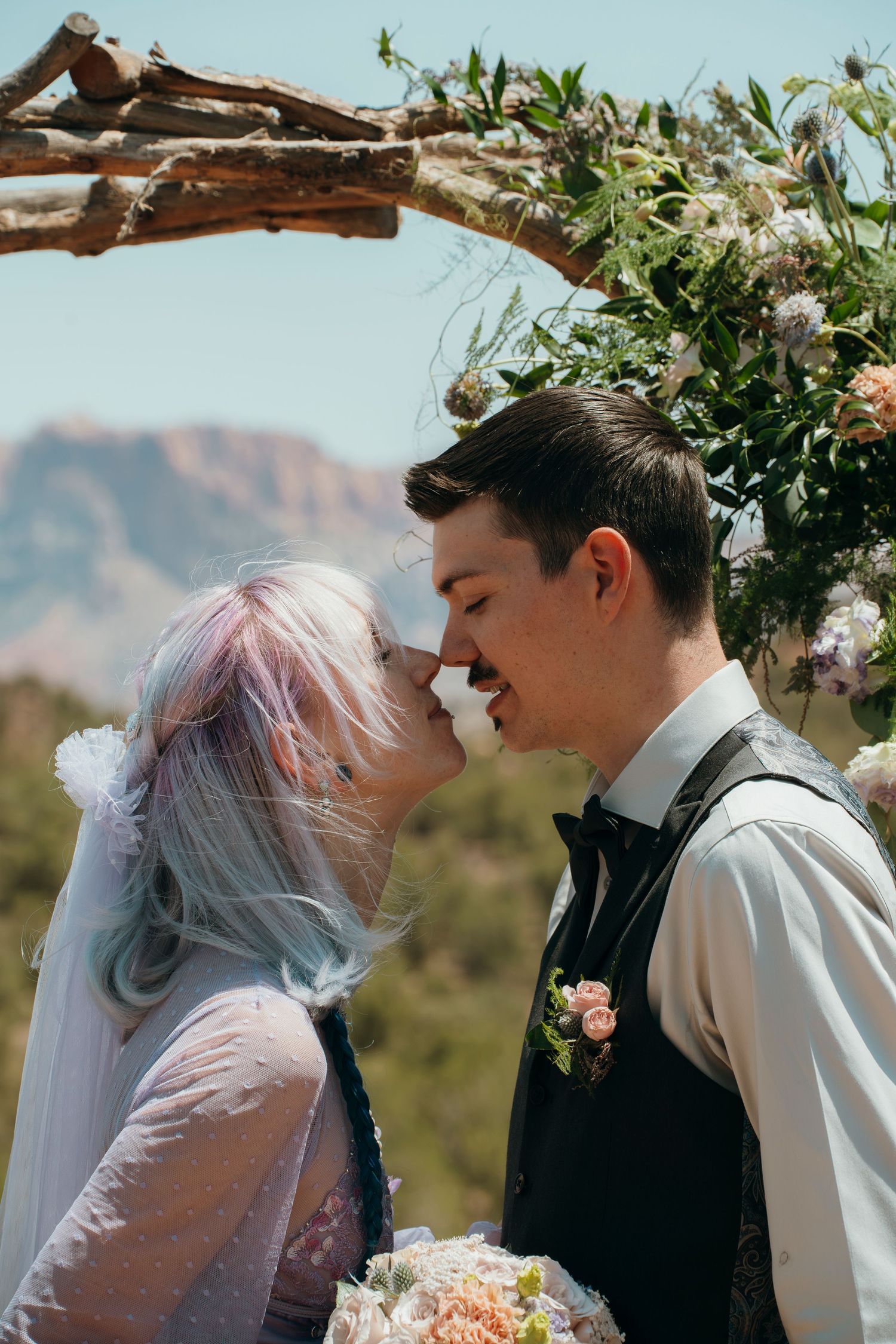 Bride with pastel hair and groom share a romantic moment under a floral arch with mountain views.