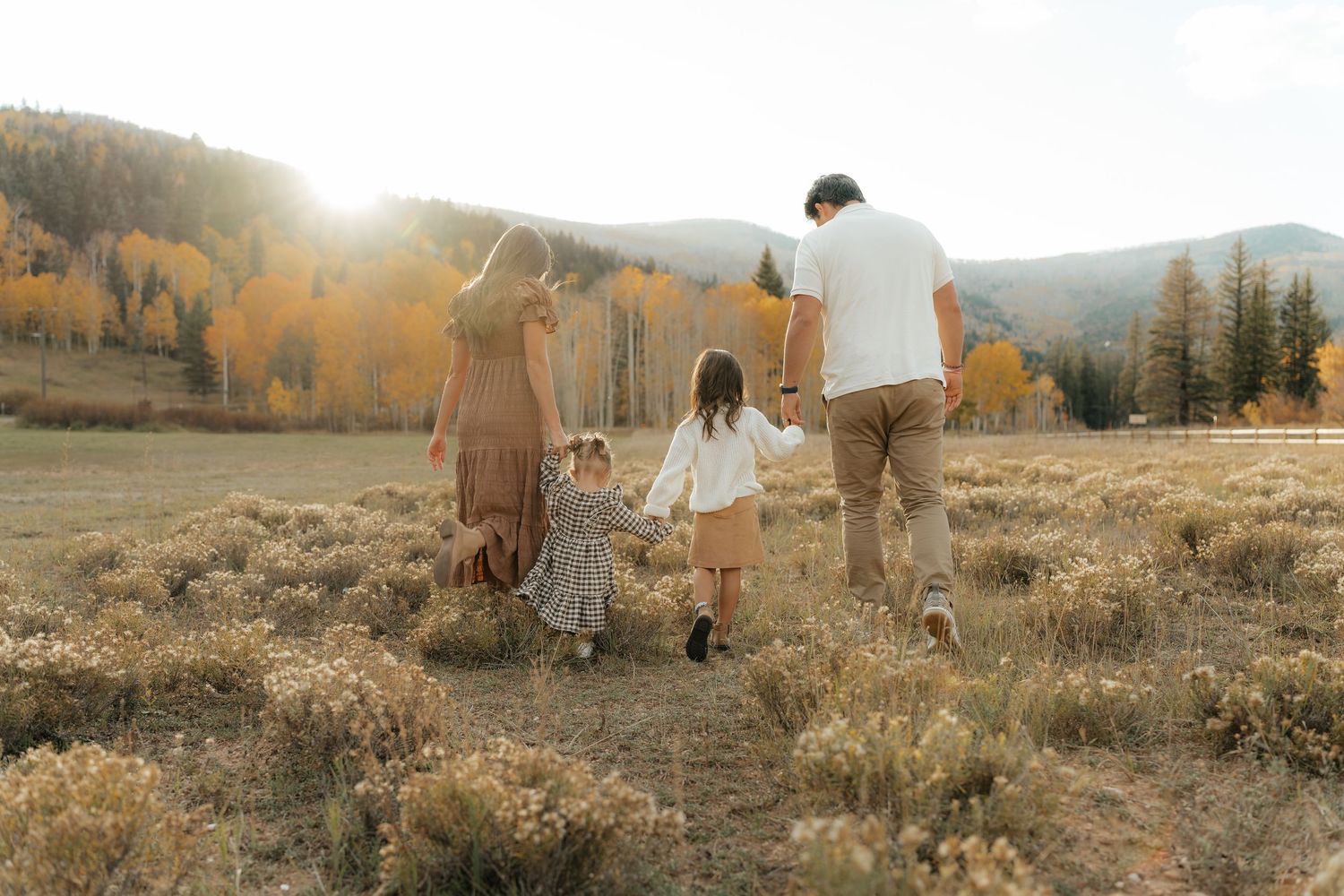 Family of four holding hands and walking away on a dirt path through a golden meadow with autumn foliage and mountains in warm sunset light.
