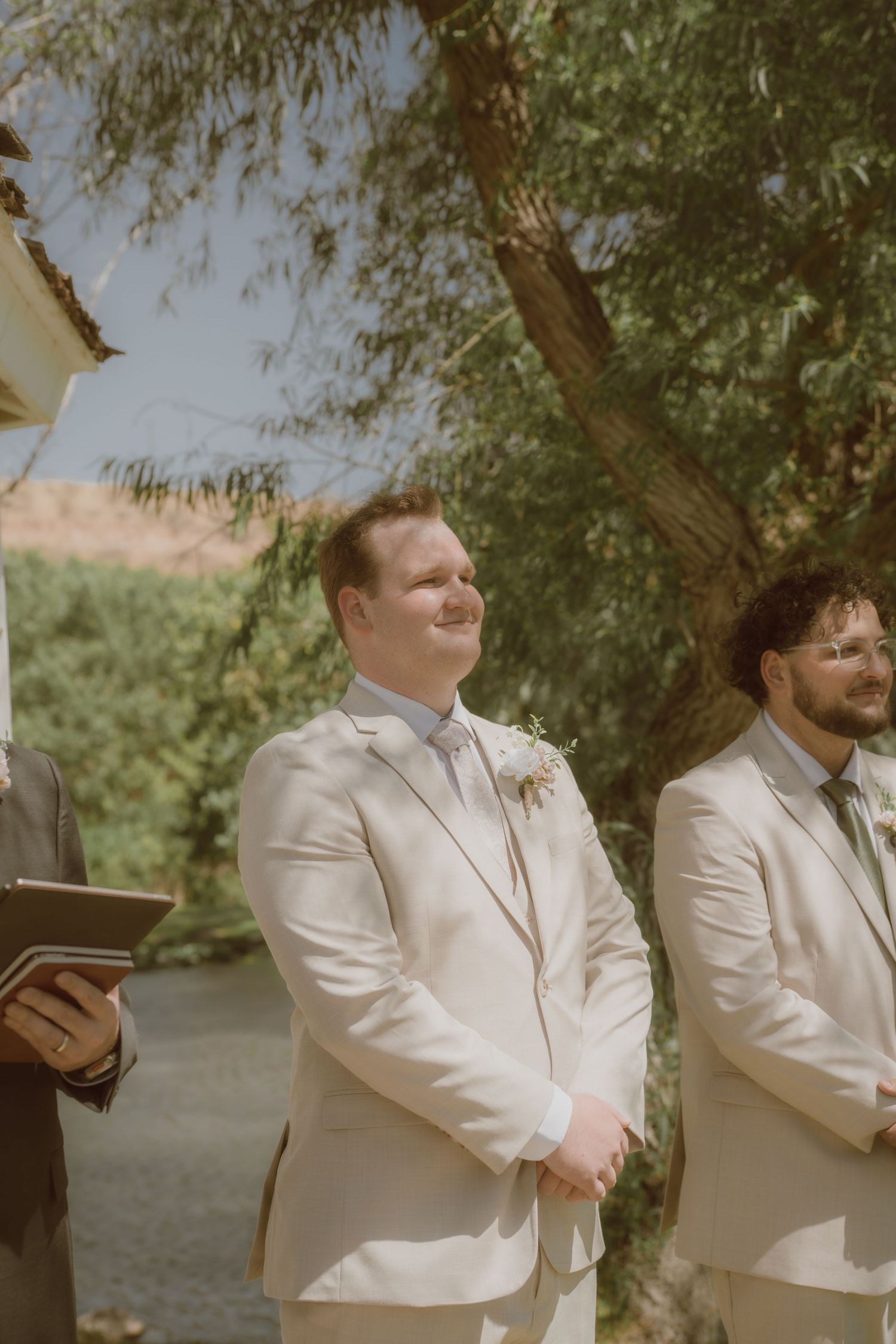 Groom in beige suit with boutonniere smiling emotionally at wedding ceremony outdoors with groomsmen standing nearby.