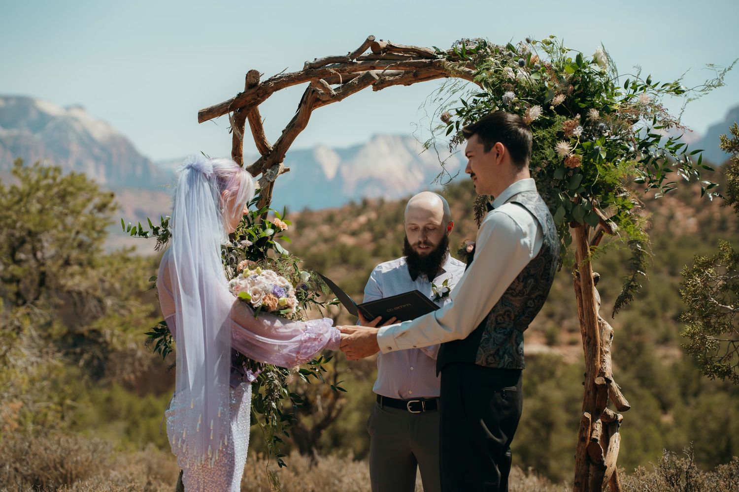 Couple exchanging vows under rustic floral arch at outdoor desert wedding with mountain backdrop.