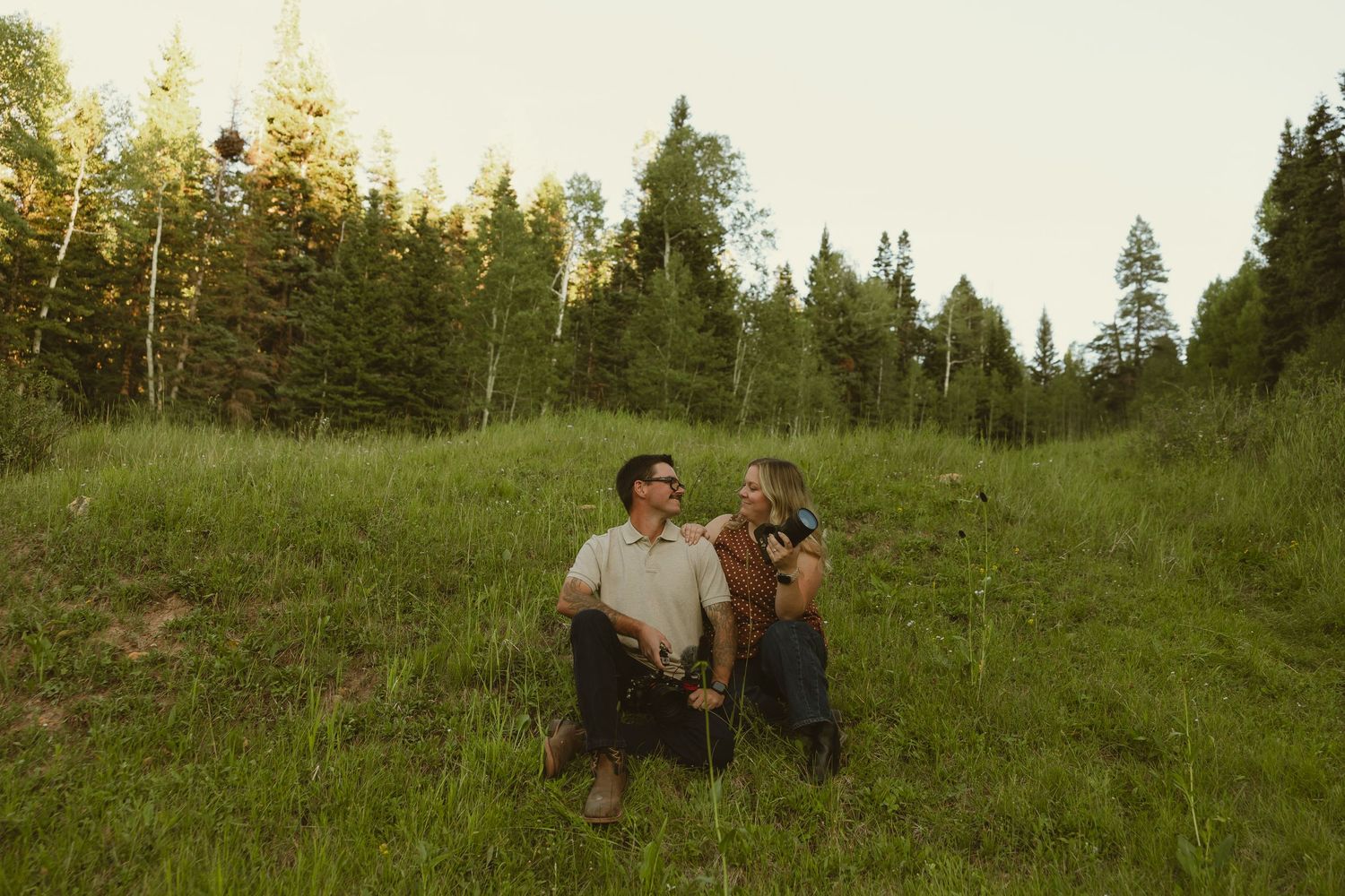 Couple sitting together in a lush green meadow surrounded by tall pine trees during golden hour.