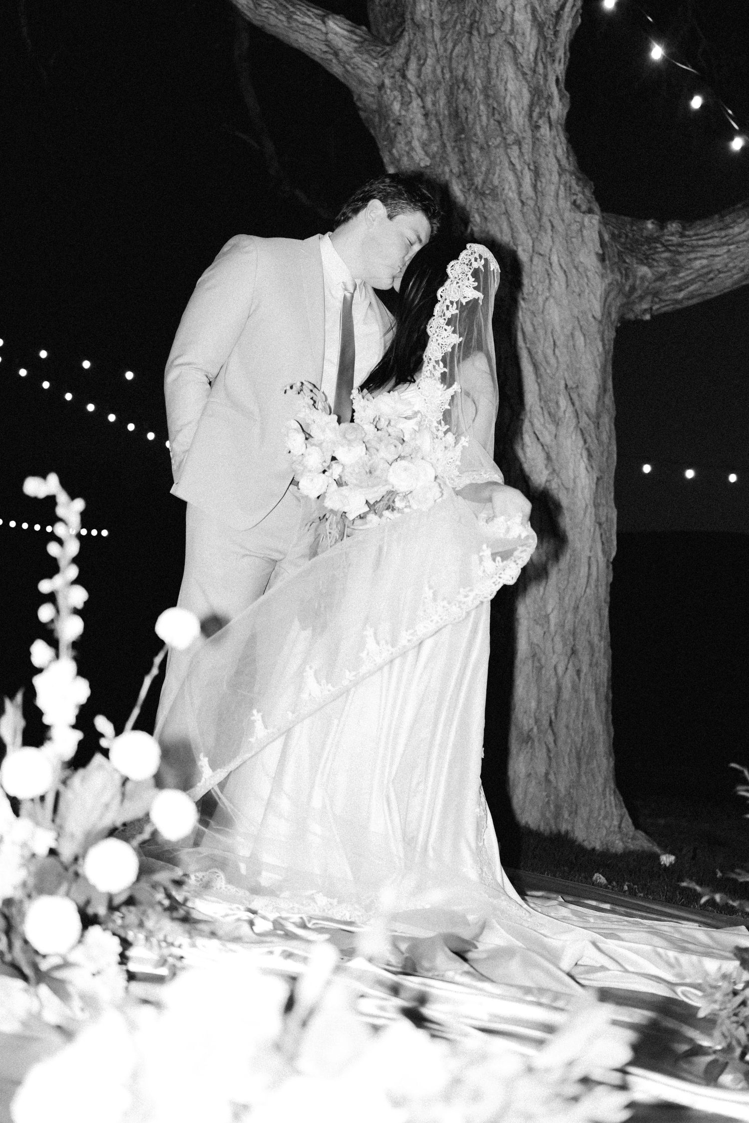 Black and white romantic wedding portrait of bride and groom kissing under a tree with string lights.