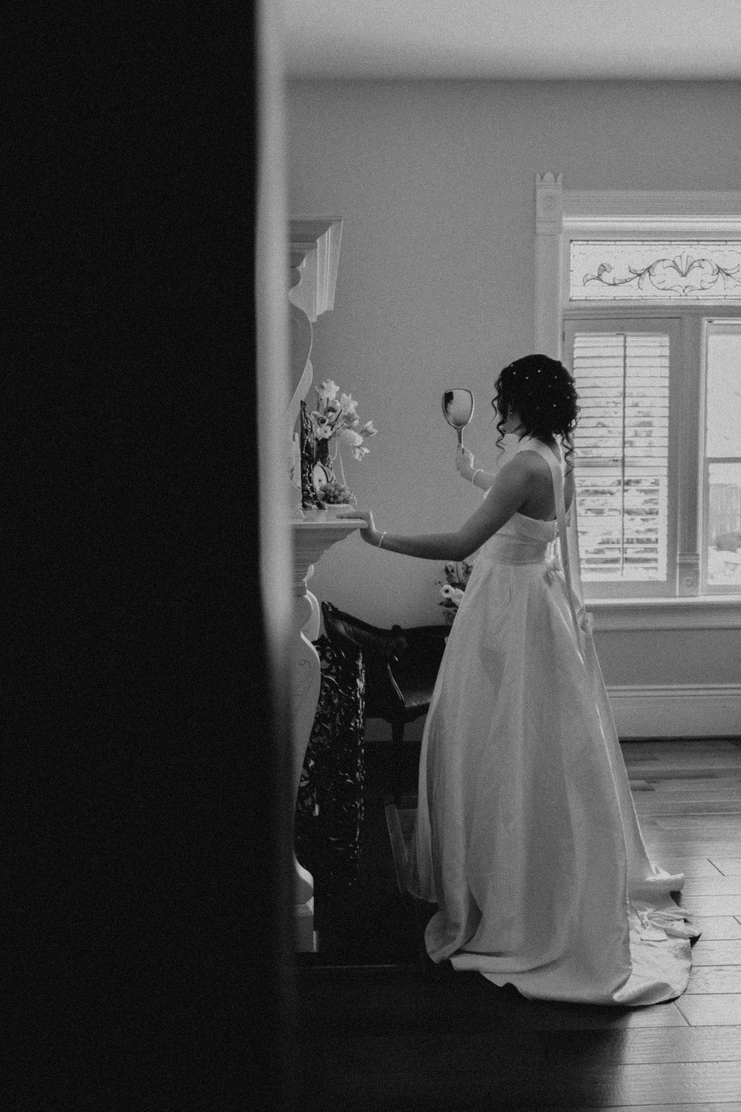 Bride in white gown holds hand mirror while getting ready in elegant room, captured in black and white.