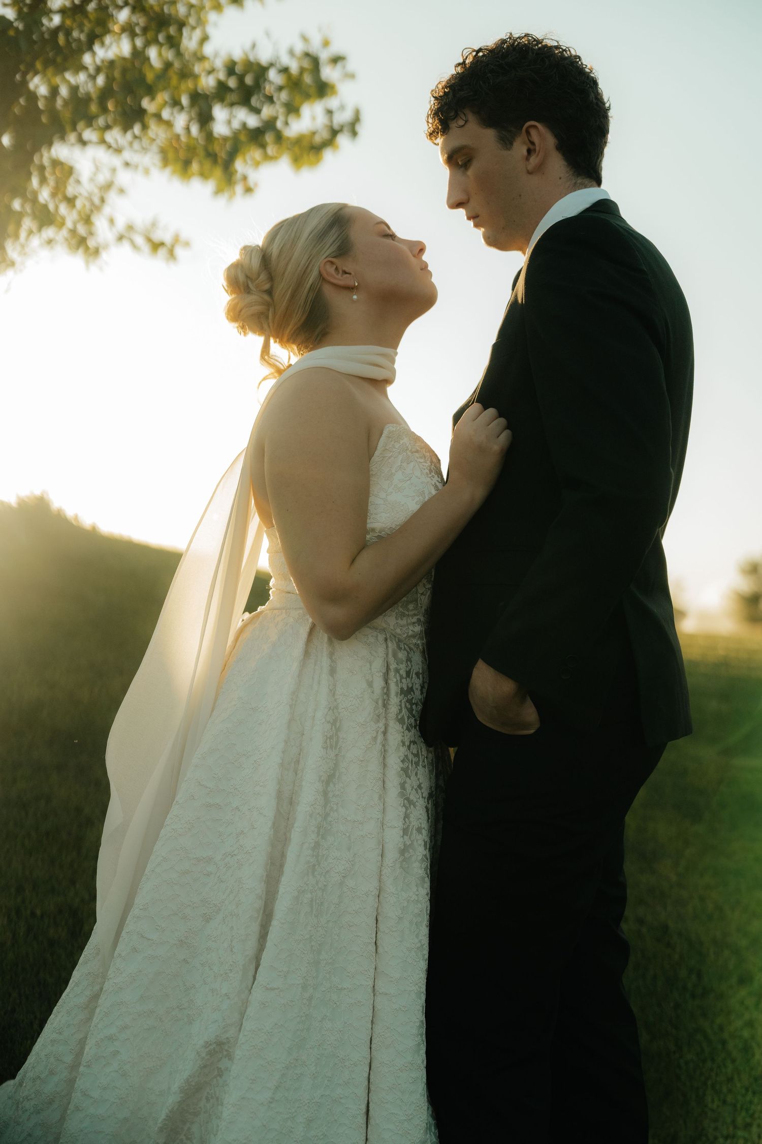 Bride and groom share romantic sunset moment outdoors, she in white halter gown with veil, he in black suit.