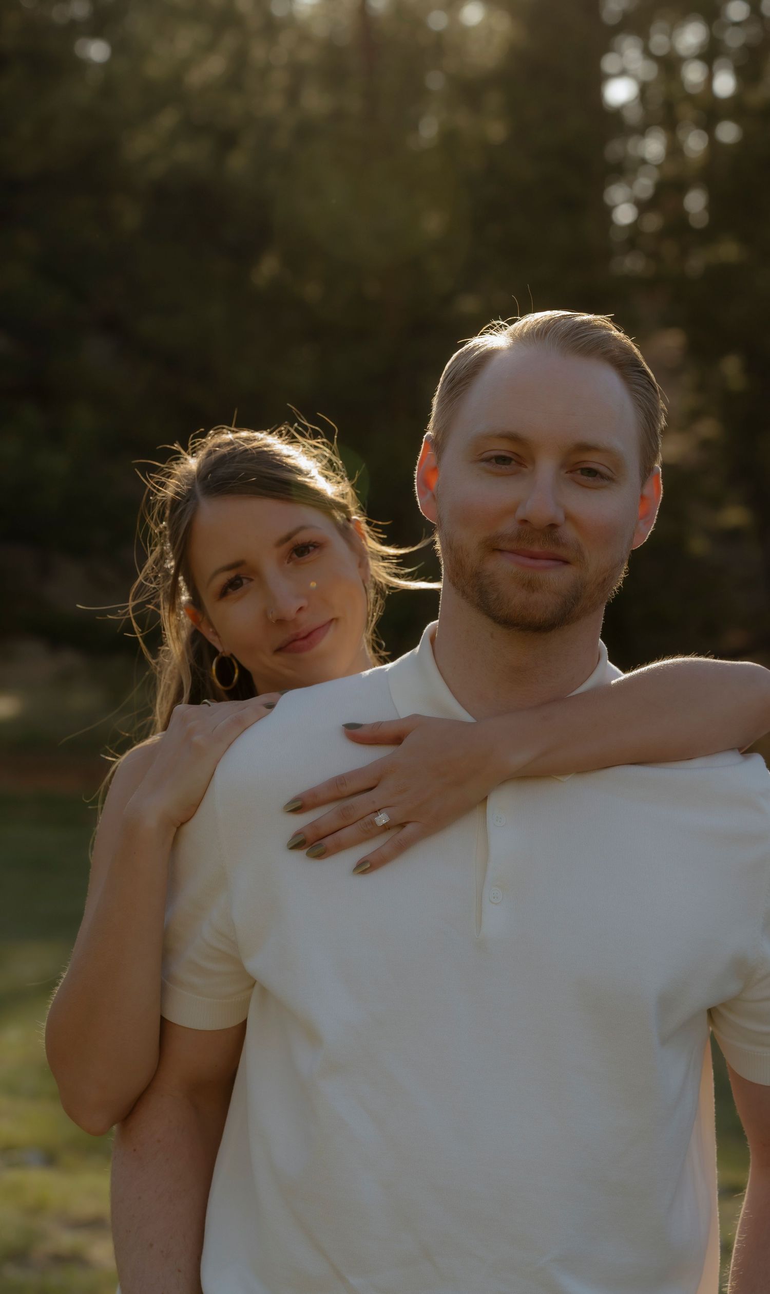 A couple in matching cream tops poses outdoors at golden hour, woman resting hands on mans shoulders.