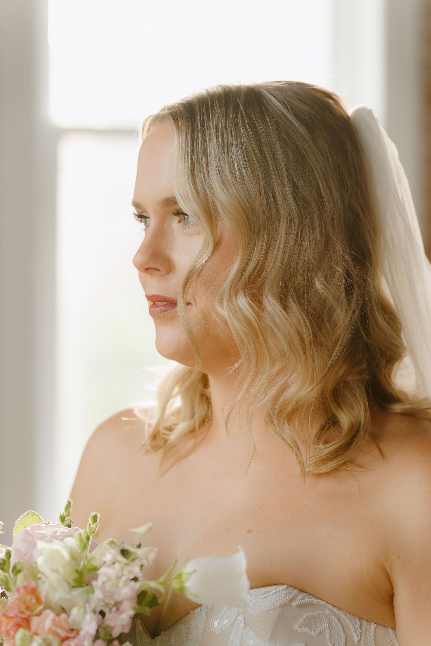 A bride with soft wavy blonde hair and a veil holds a pastel bouquet, gazing thoughtfully toward a bright window.