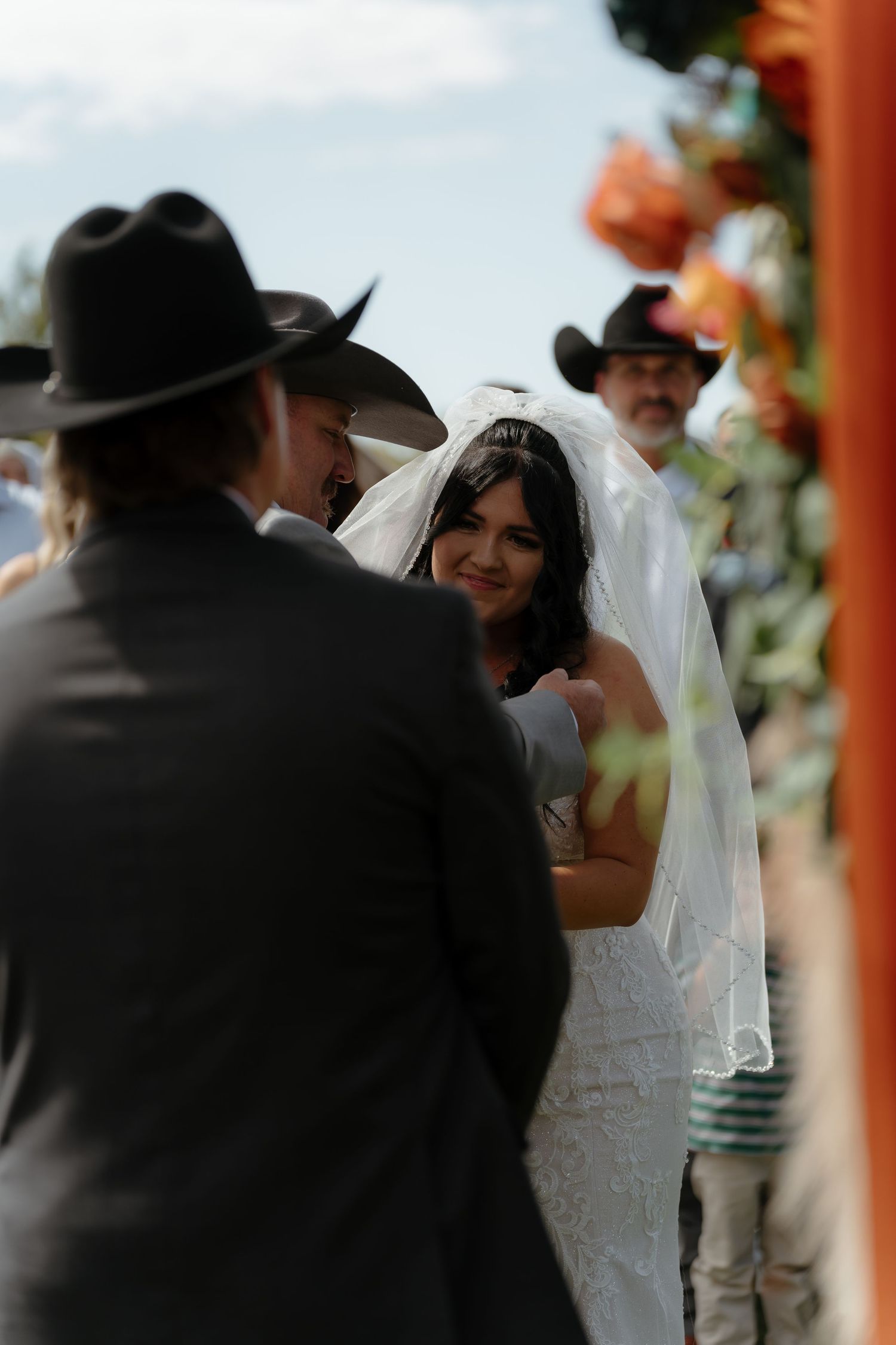 Smiling bride in white lace gown and veil exchanges vows with groom in black cowboy hat at outdoor wedding.