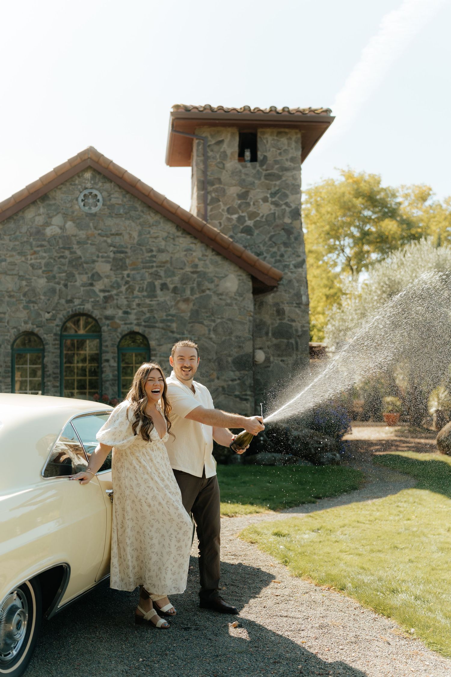 Laughing couple pops champagne beside a vintage cream car in front of a rustic stone building with a tower on a sunny day.