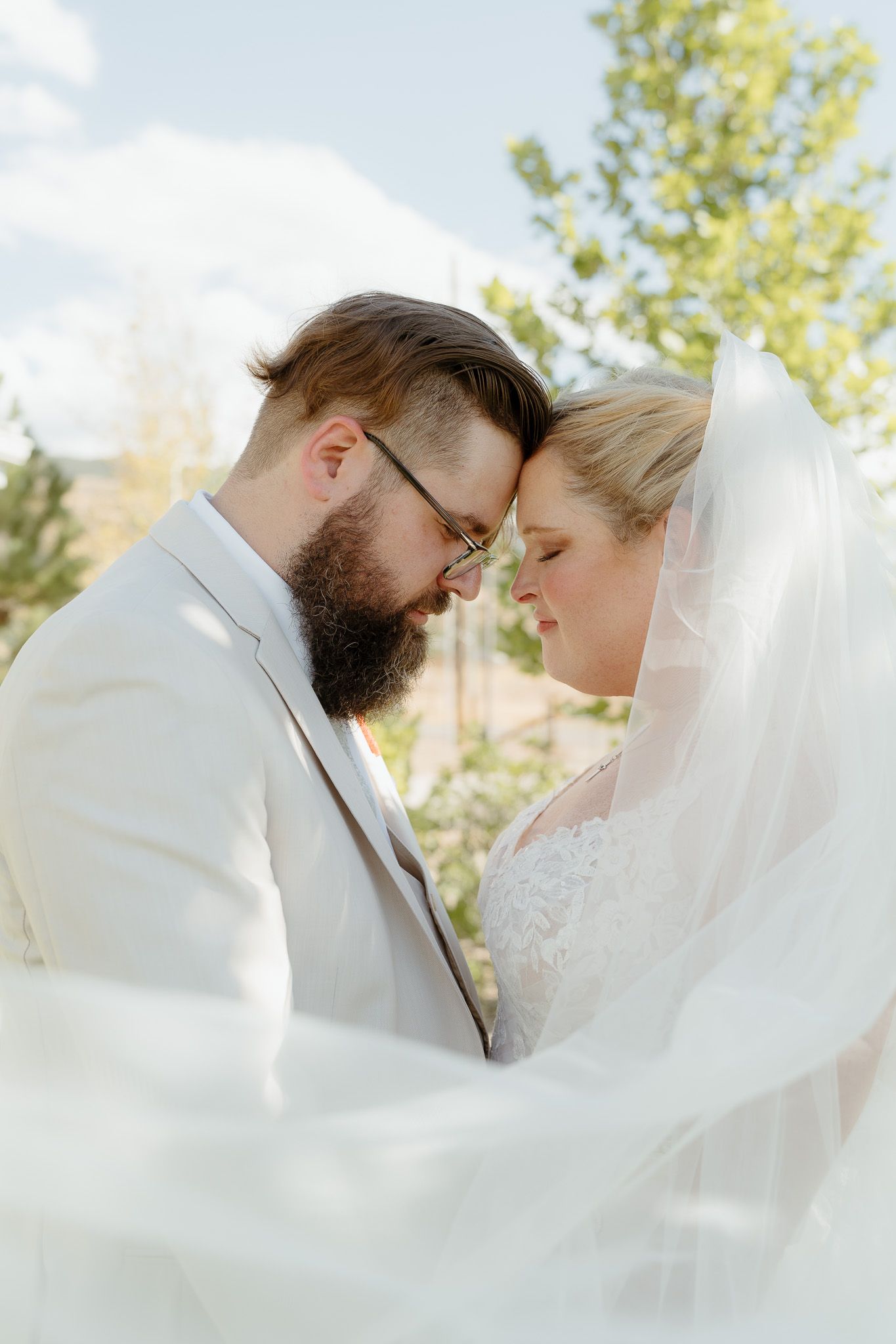 Bride and groom touch foreheads tenderly under a flowing white veil outdoors on their wedding day.
