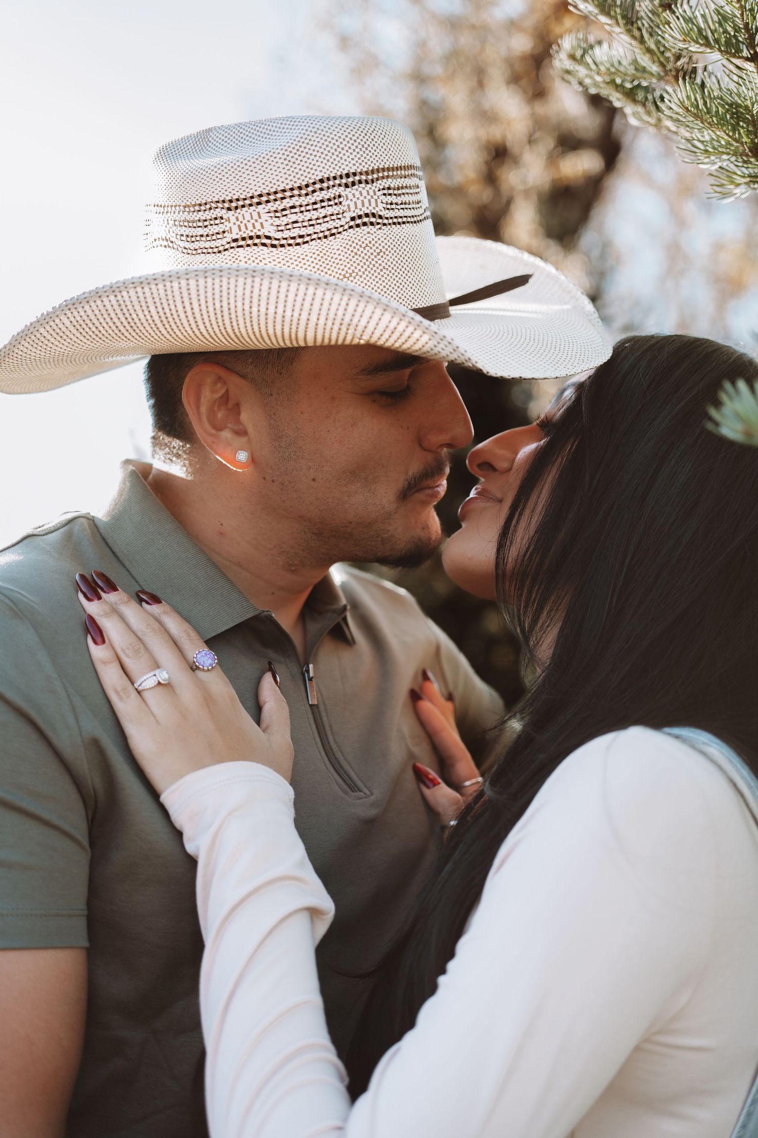 Couple wearing matching cowboy hats share a kiss outdoors in warm sunlight during engagement session.
