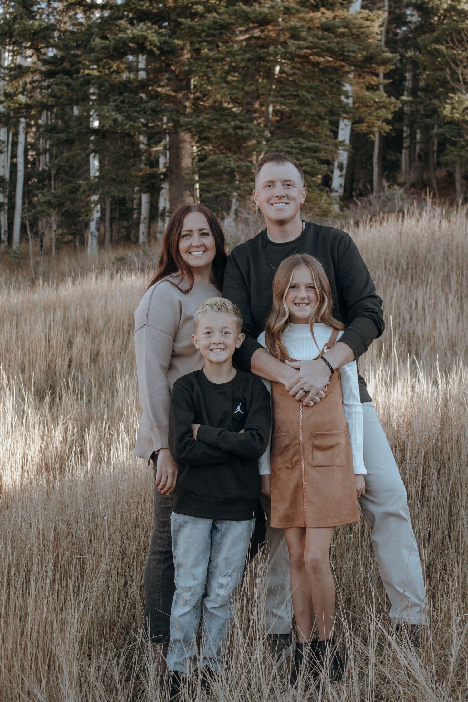 Family of four posing in golden field with forest backdrop during autumn outdoor photo session.