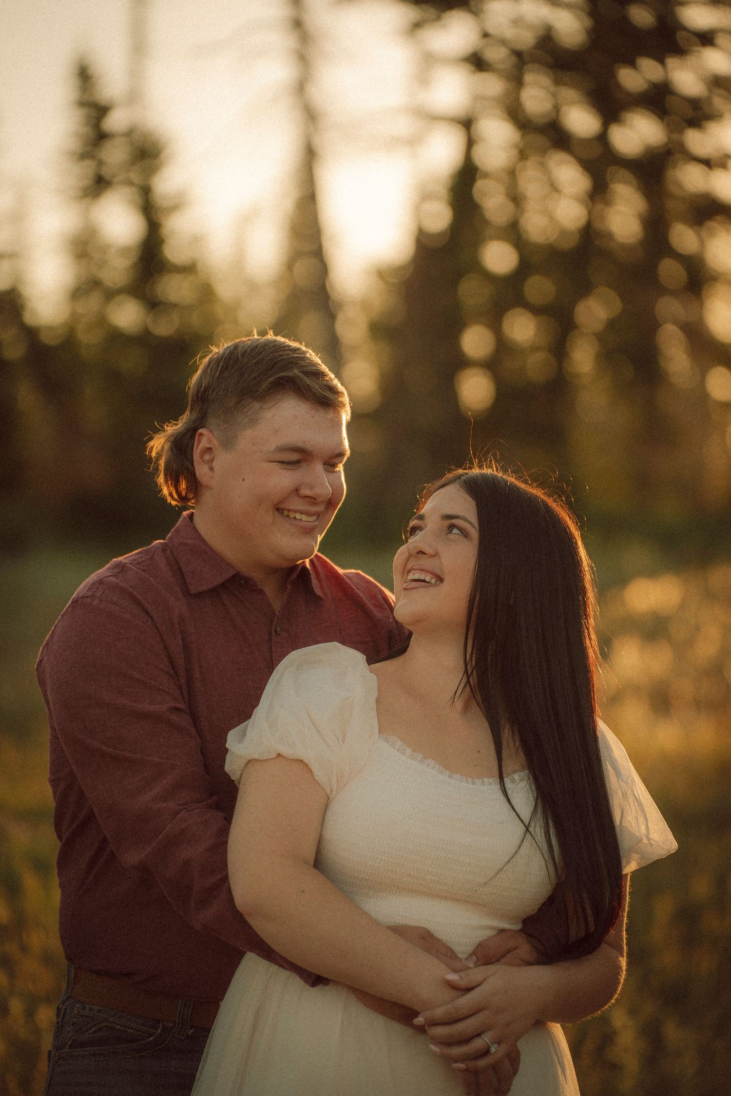 Couple embracing at golden hour in a forest, smiling at each other, woman in white dress, man in red shirt.
