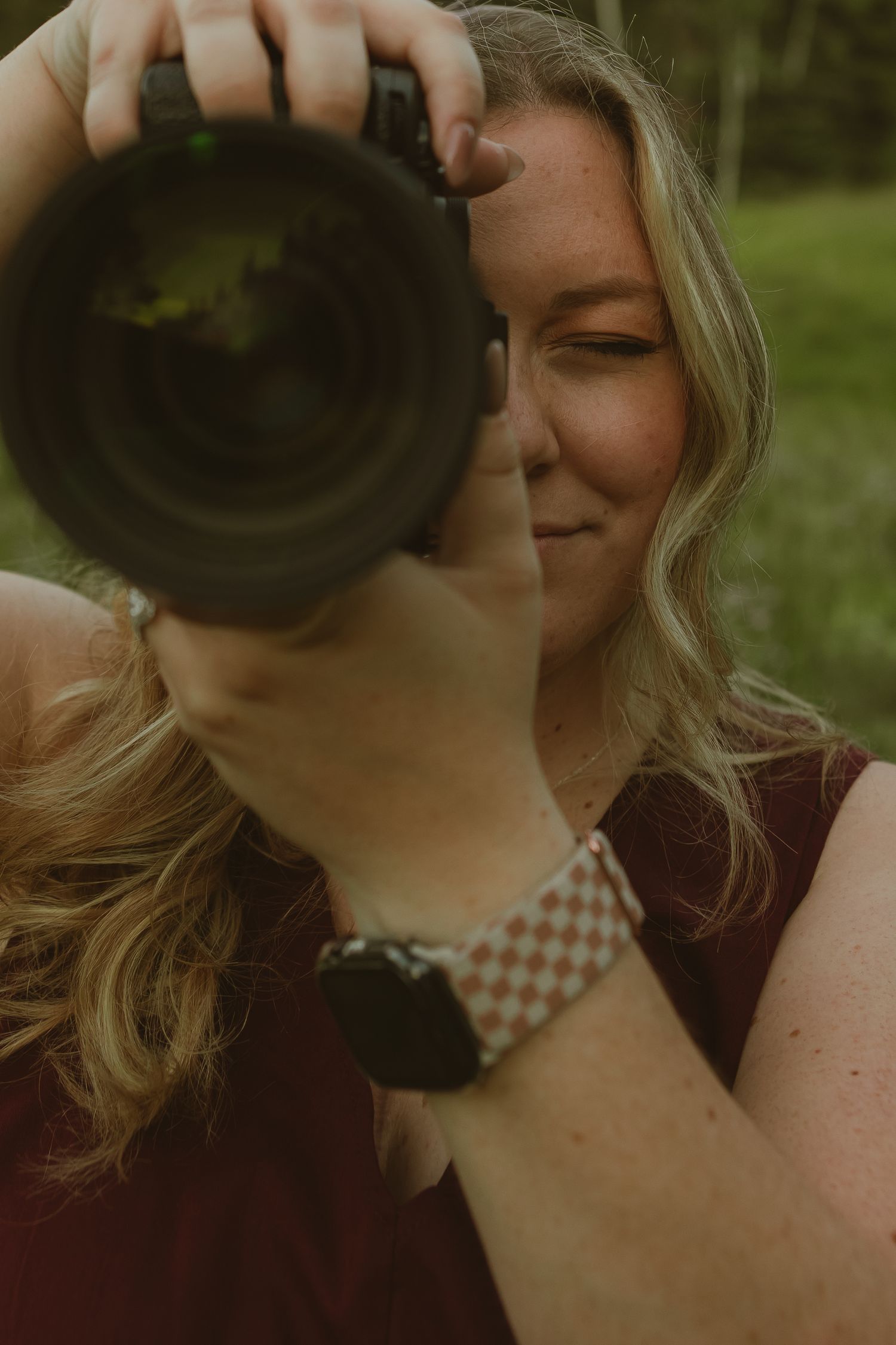 A blonde woman in a burgundy top photographs with a DSLR camera, wearing a checkered watch band outdoors.