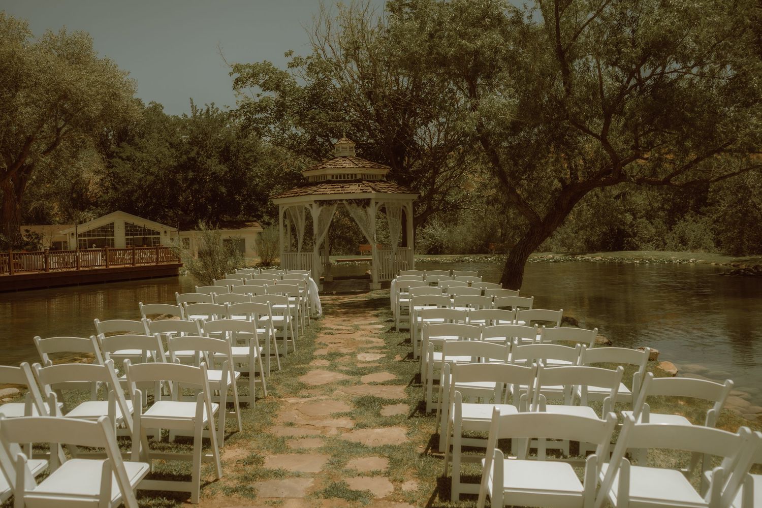 Outdoor wedding ceremony setup with white chairs along a stone path leading to a wooden gazebo surrounded by lush trees by a river.