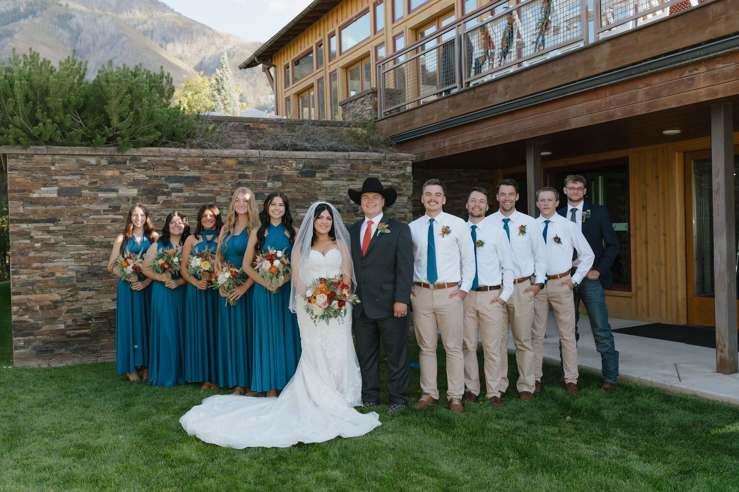 Wedding party in teal dresses and khaki suits poses outdoors by stone building with mountain backdrop.