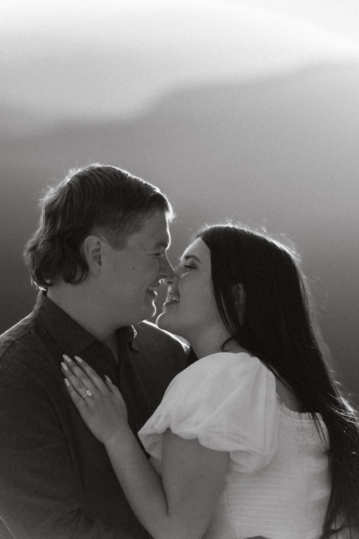 Black and white photo of a couple about to kiss, woman wearing engagement ring, soft backlit mountain scenery.