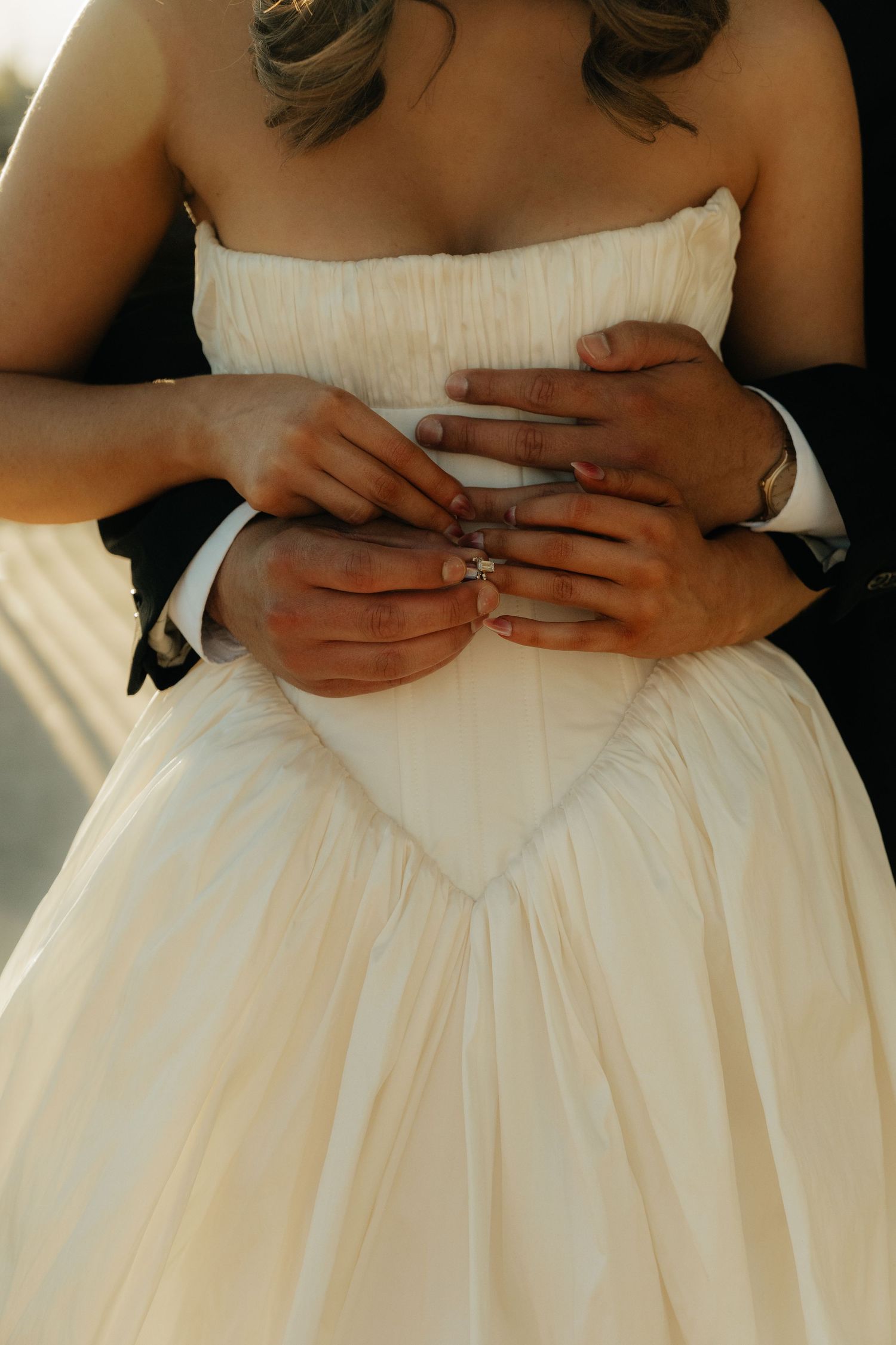 Bride and groom holding hands showing wedding rings, bride wearing elegant strapless white gown.