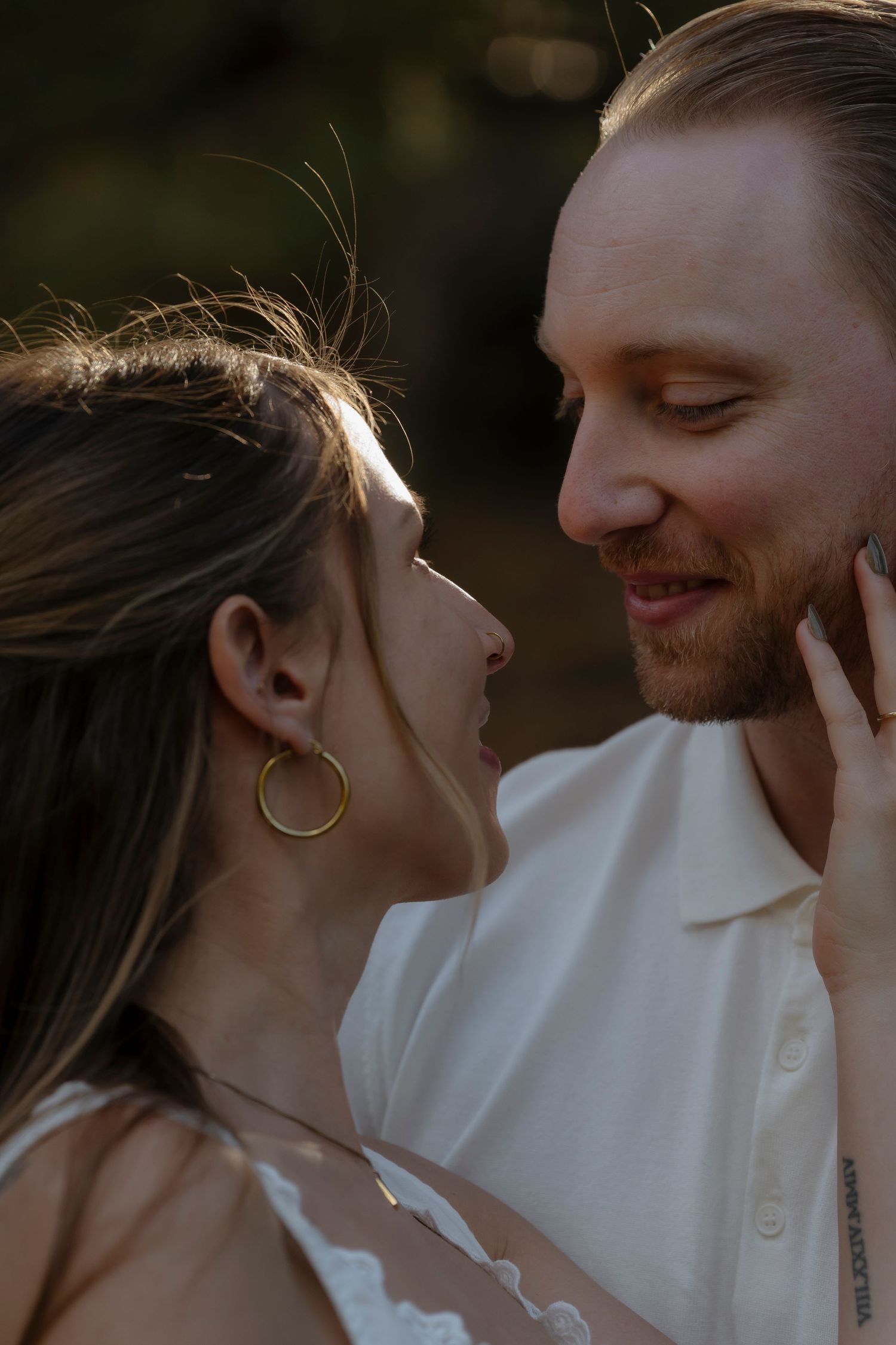 A couple shares a romantic close moment, the woman wearing gold hoop earrings and the man smiling in a white shirt.