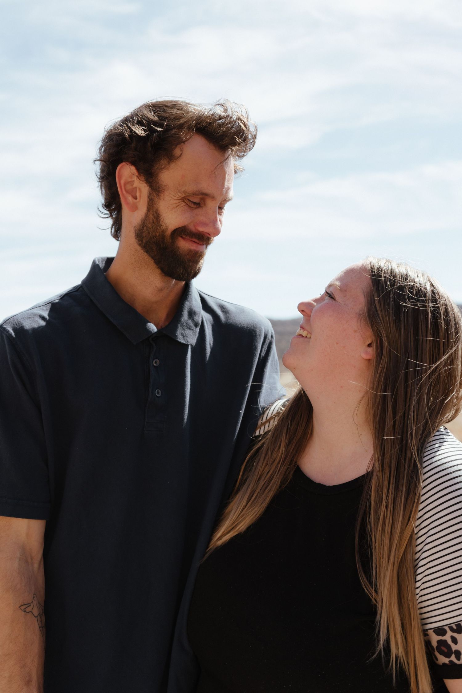 A smiling couple gazing at each other outdoors under a blue sky, man in black polo and woman with long hair.
