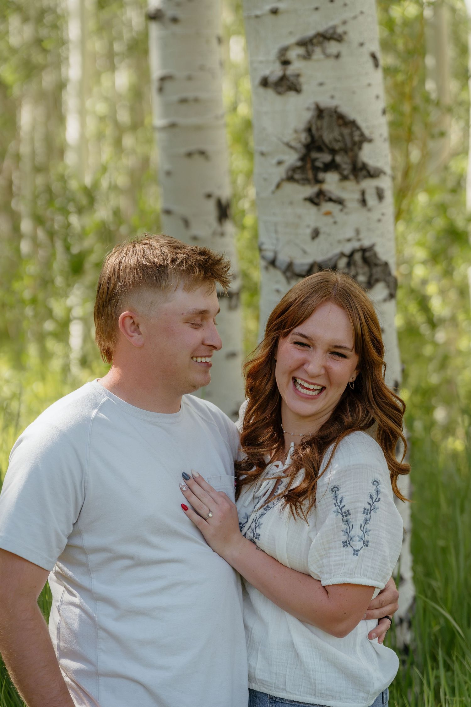 A laughing couple embraces among white birch trees in a sunny forest during an outdoor engagement photo session.