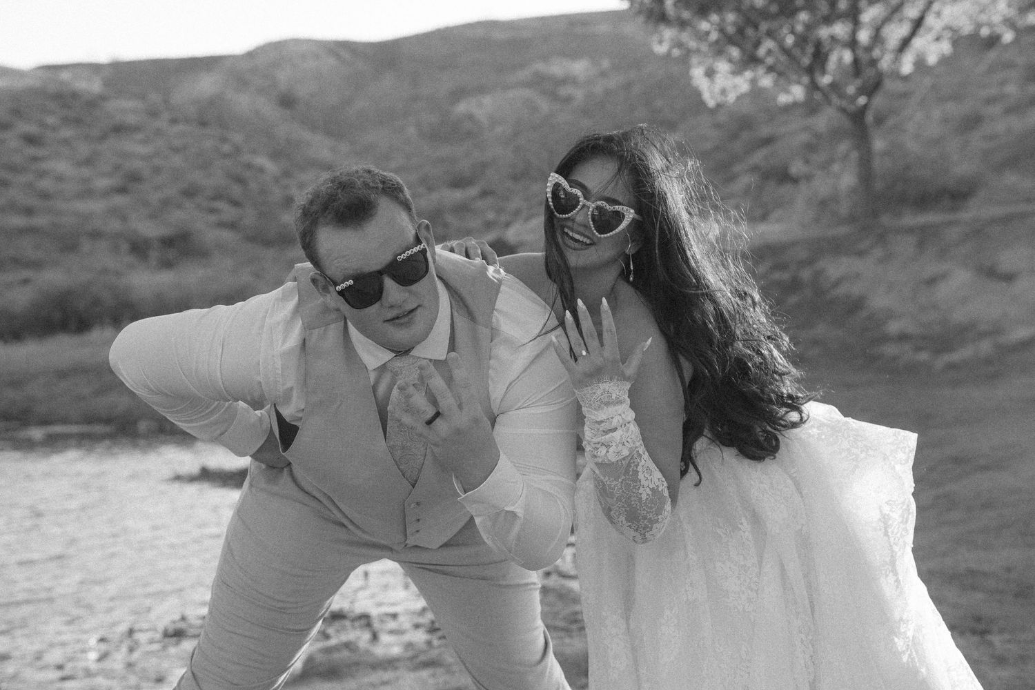 Black and white photo of a bride and groom in sunglasses striking fun poses outdoors near a river.