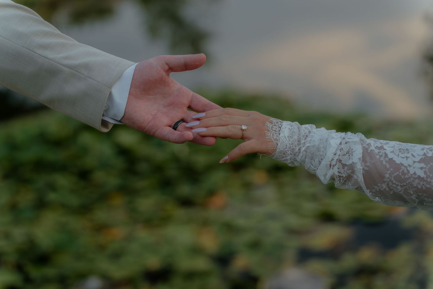 Bride and groom holding hands, wedding ring visible on bride's finger, lace sleeve, romantic outdoor ceremony.
